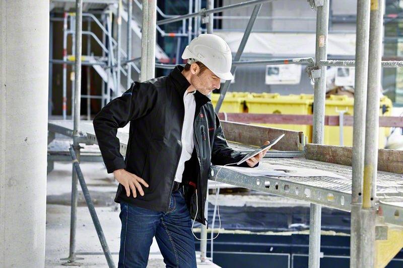 Muškarac stoji na gradilištu, nosi zaštitnu kacigu i promatra tablet. U pozadini se vide skele.