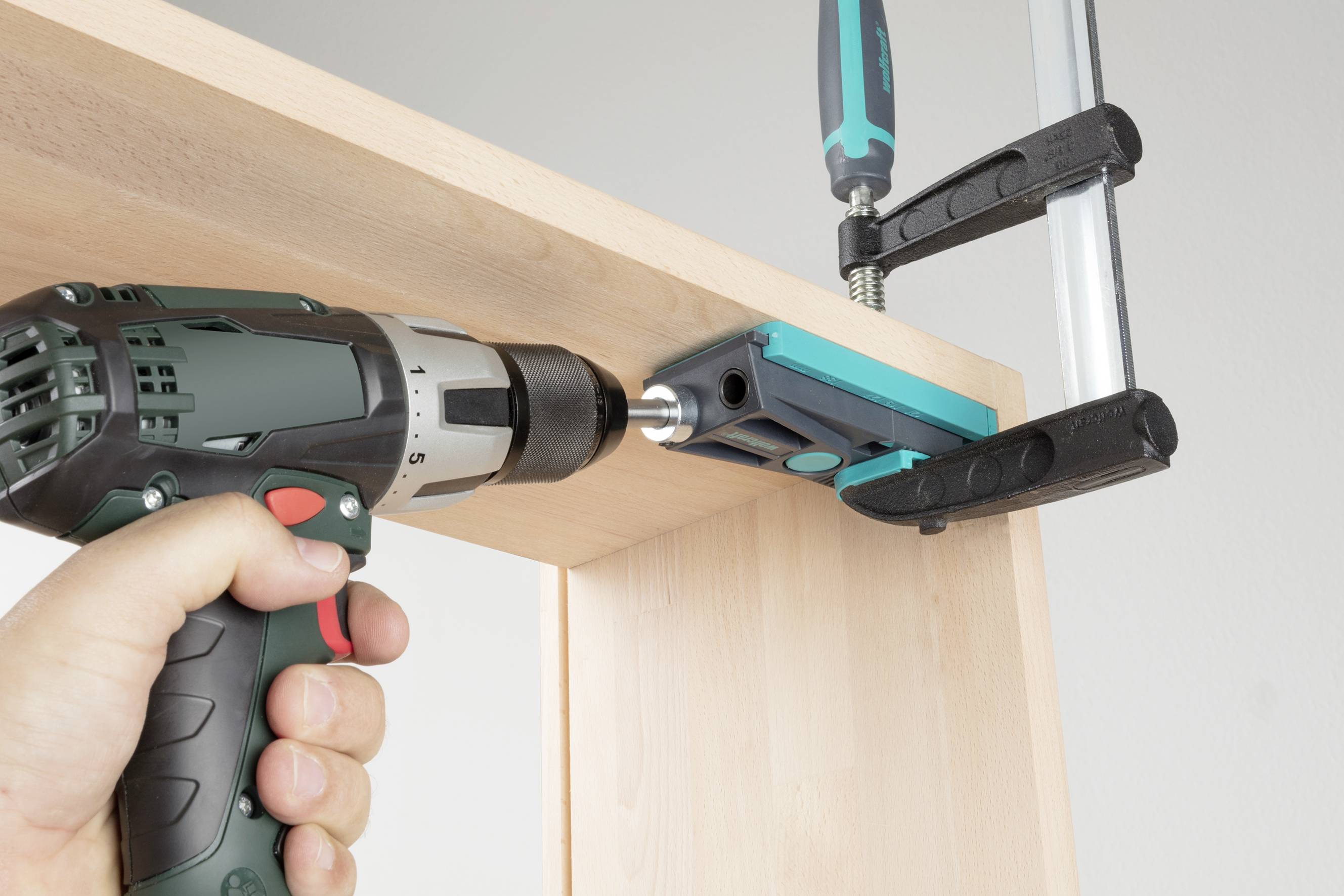 Bušilica se koristi za sastavljanje drvene police, potpomognuta stegom. Ruka čvrsto drži bušilicu.