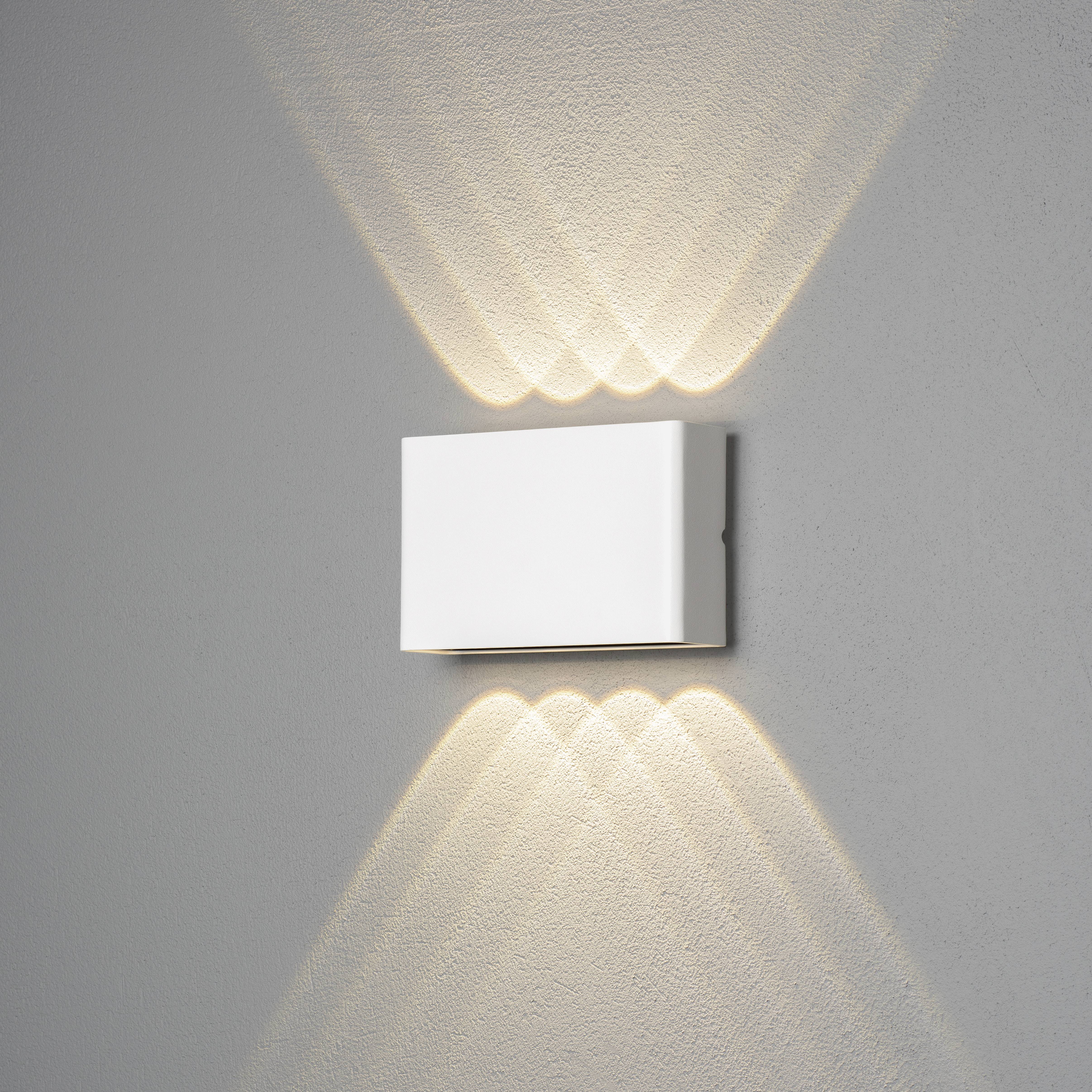Zidna lampa zrači toplim svjetlom u dekorativnom uzorku prema gore i dolje, montirana na sivoj stijeni. Minimalistički dizajn.
