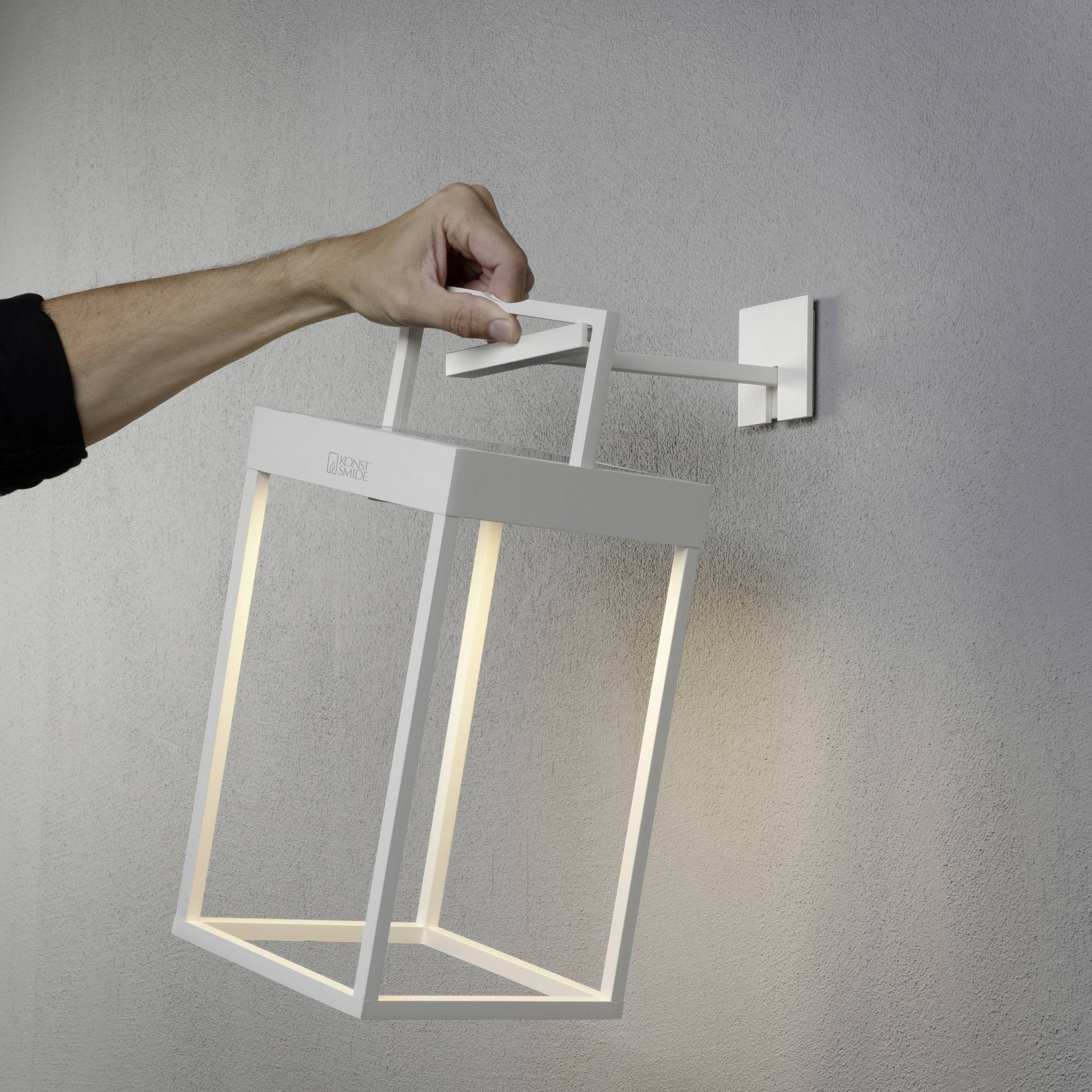 Ruka montira modernu, minimalističku, bijelu zidnu lampu na sivoj stijeni. Lampa ima pravokutan, otvoren oblik.