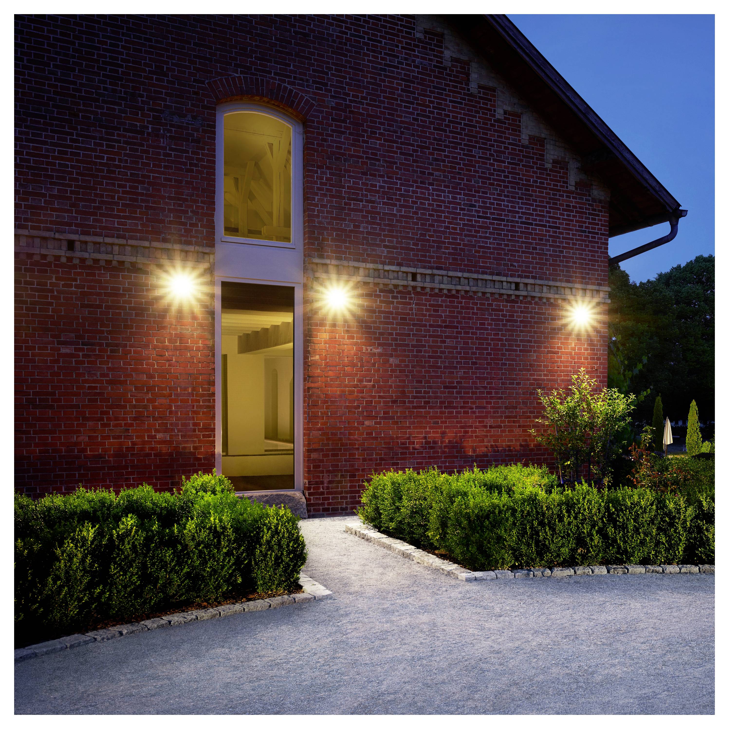 Kuća od opeke sa vanjskom stranom na sumrak s osvijetljenim ulazom, tri zidna svjetla i urednim živicama porezanim duž šljunčane staze.