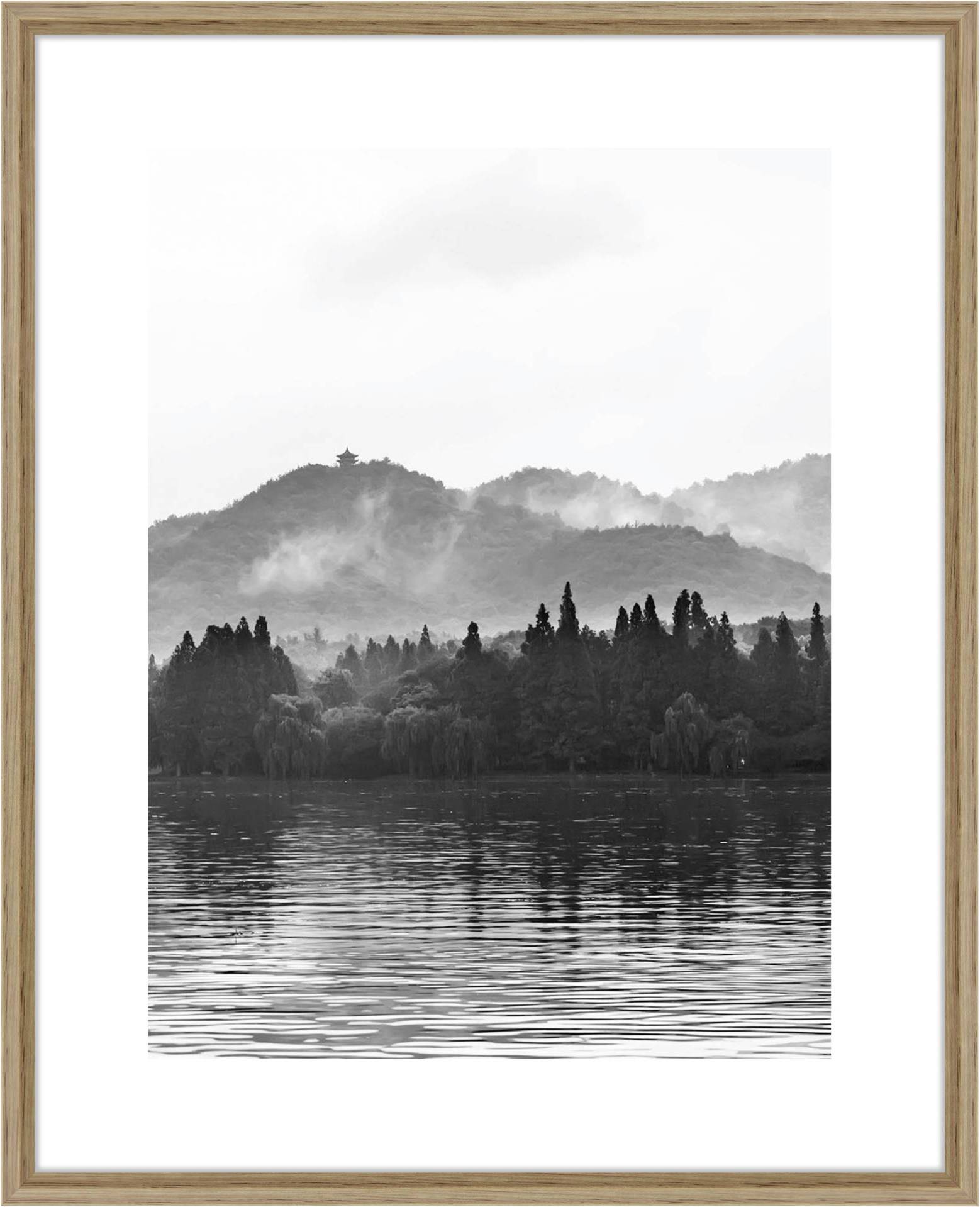 Crno-bijela fotografija tihog jezera sa stablima i brdima u pozadini, maglovito raspoloženje, mirni krajolik.