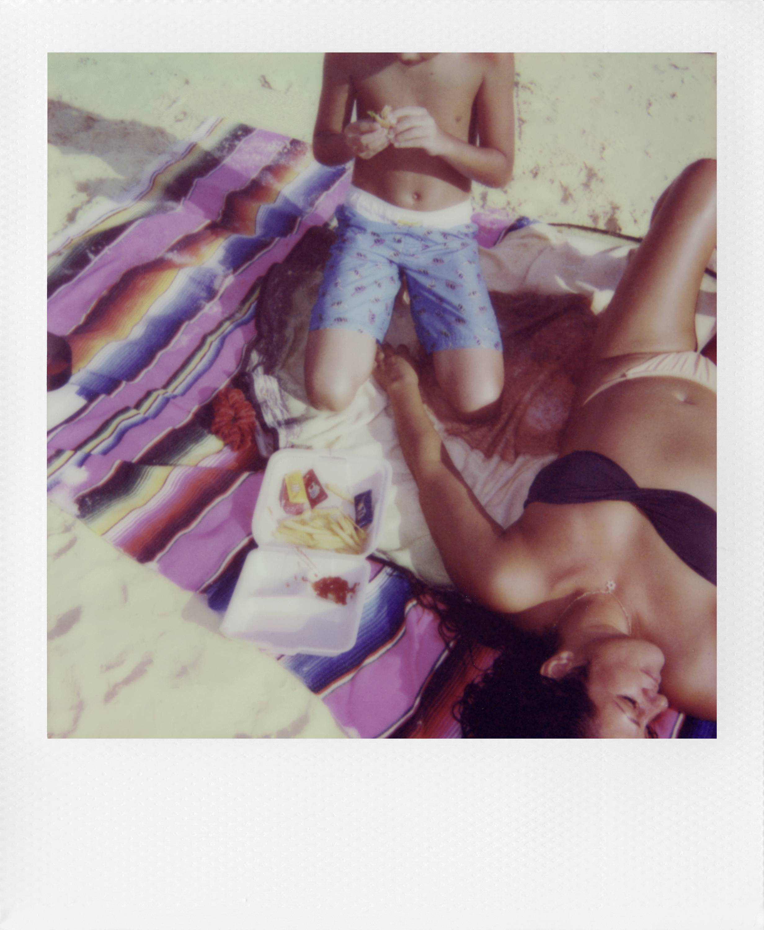 Žena u bikiniju leži na šarenoj deki na plaži, dok dijete u kratkim hlačama stoji pored nje i drži nešto.