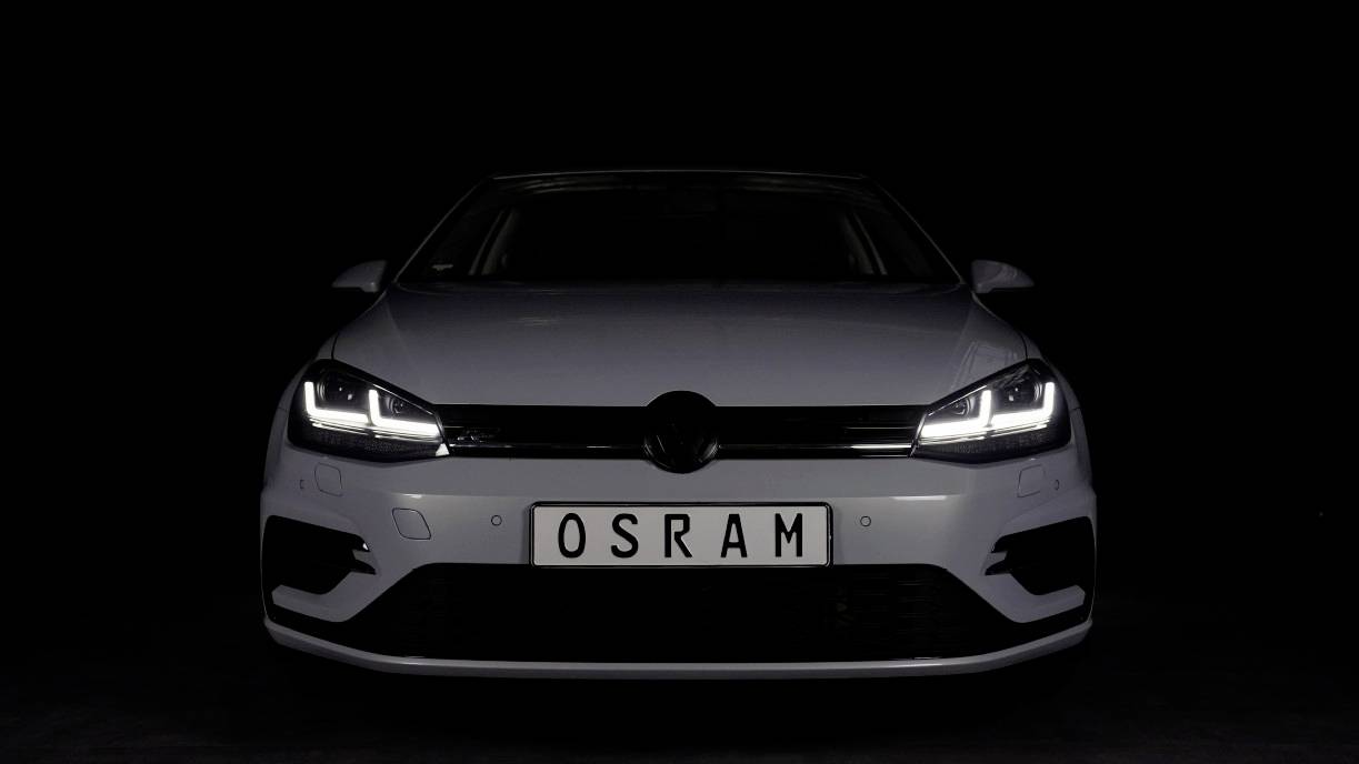 Bijeli automobil s uključenim LED dnevnim svjetlom, frontalno snimljen ispred crne pozadine. Na registarskoj tablici piše 'OSRAM'.
