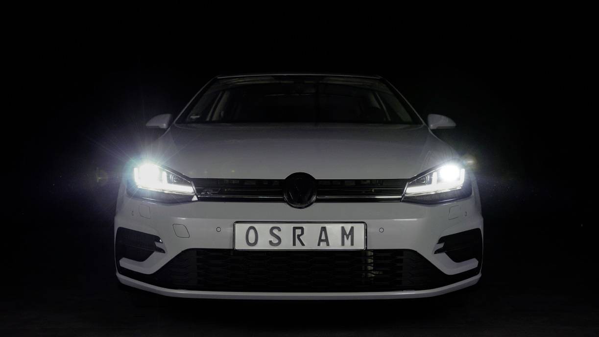 Bijeli automobil s uključenim farovima prikazuje registarsku tablicu 'OSRAM' pred tamnom pozadinom.