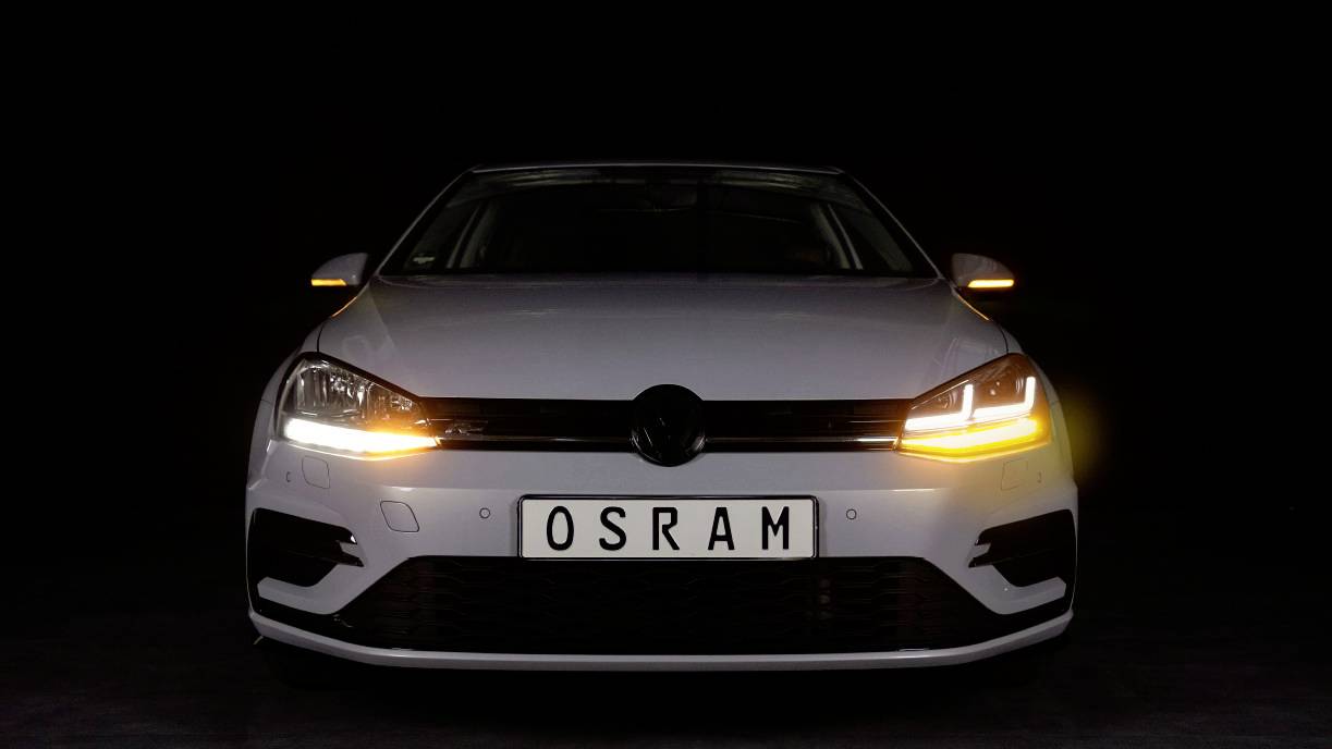 Bijeli automobil s osvjetljenim farovima i registarskom tablom na kojoj piše 'OSRAM' stoji ispred tamne pozadine.