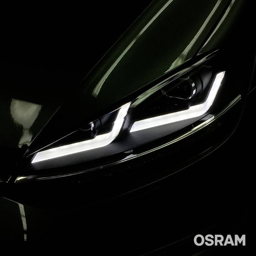 Bliski kadar prednjeg automobilskog farova s modernim, svijetlećim LED-trakama. Logo 'OSRAM' u donjem desnom kutu. Tamna pozadina.