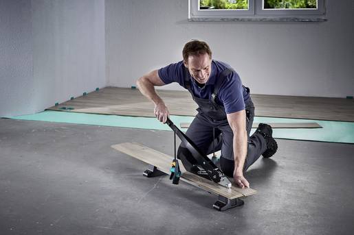 Osoba kleči na podu i koristi alat da prereže drvenu dasku. U pozadini se vidi prozor.