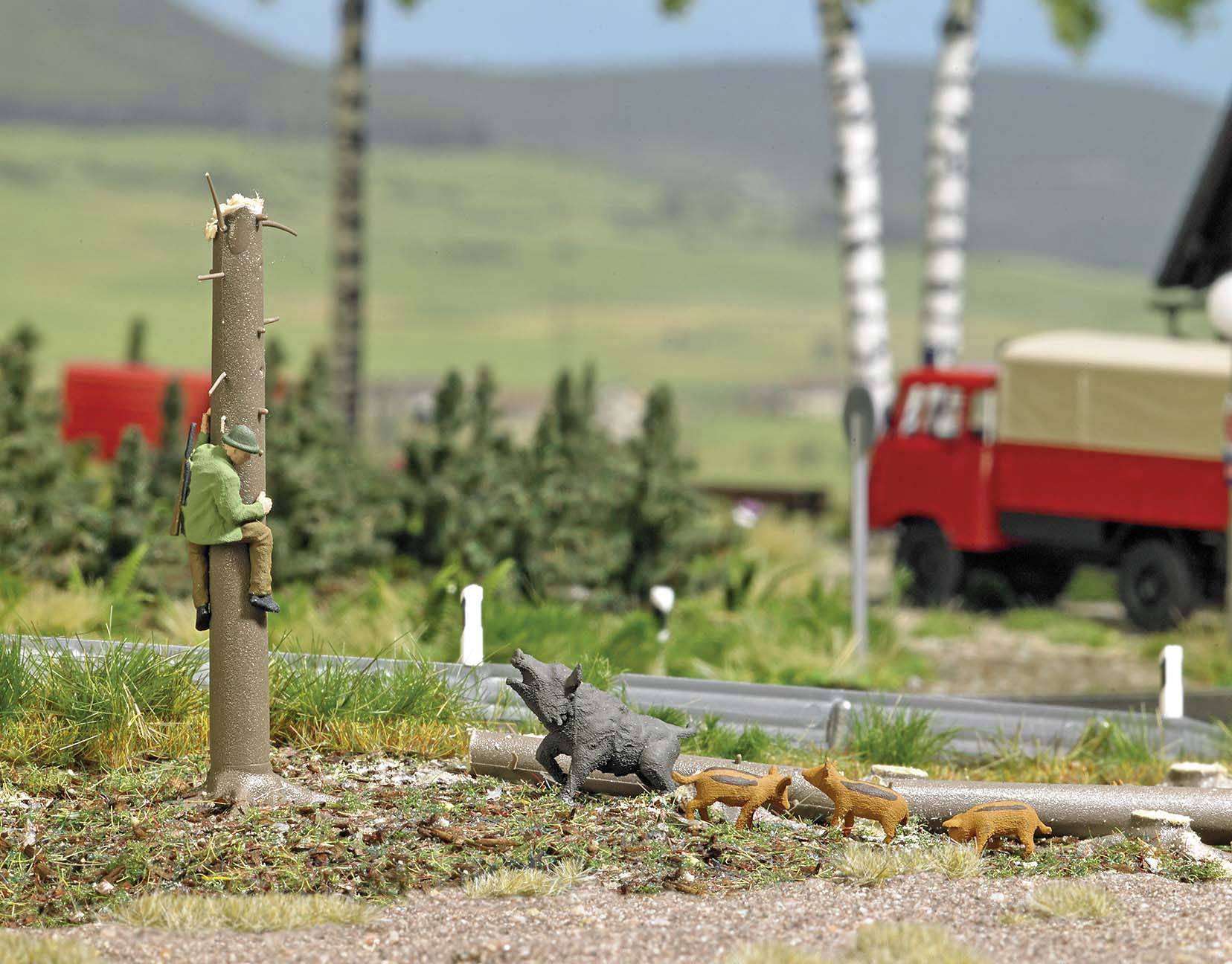 Lovac na modele penje se na drvo dok minijaturne divlje svinje i lisice šetaju oko njega. U pozadini se vide drveće i crveno vozilo.