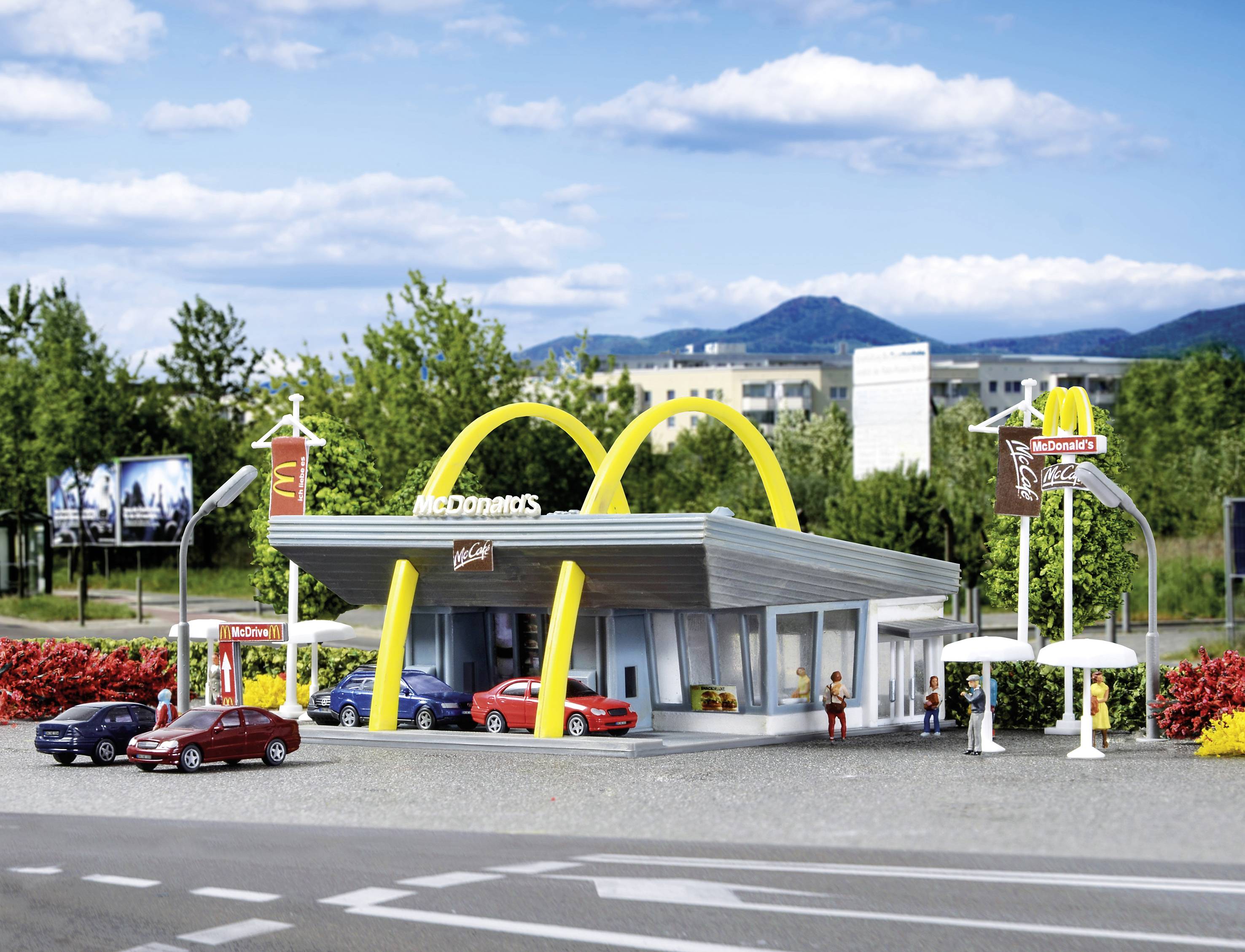 Model McDonald's restorana s tri automobila i figurama ispred, okružen drvećem i planinama u pozadini.