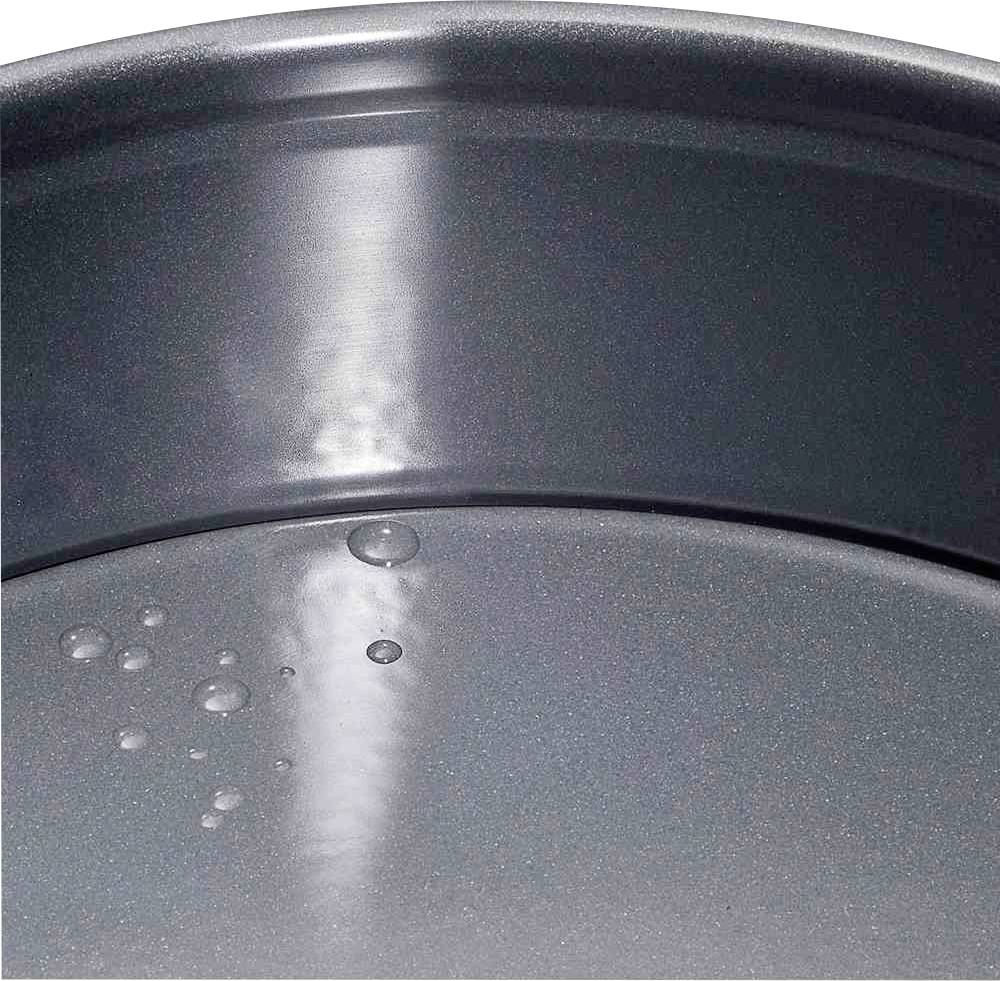 Bliski kadar praznog, okruglog aluminijskog pekača s neprianjajućim premazom. Nekoliko vodenih kapi vidljivo je na površini.