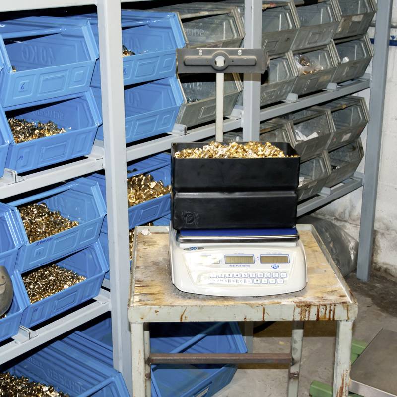 Metalni dijelovi u plavim kutijama na policama. U prvom planu: vaga s napunjenim plastičnim kontejnerom. Radi u radionici ili skladišnom prostoru.