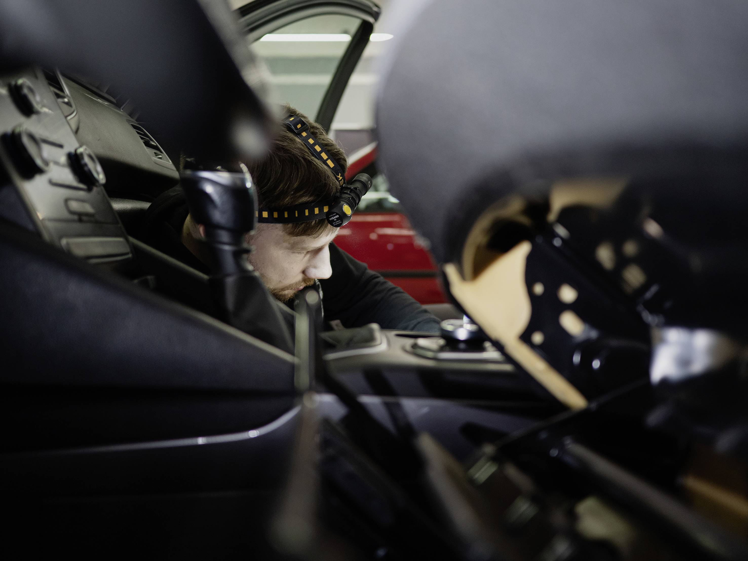 Osoba radi na unutrašnjoj opremi automobila koristeći čeono svjetlo. Crveni automobil je djelomično vidljiv u pozadini.