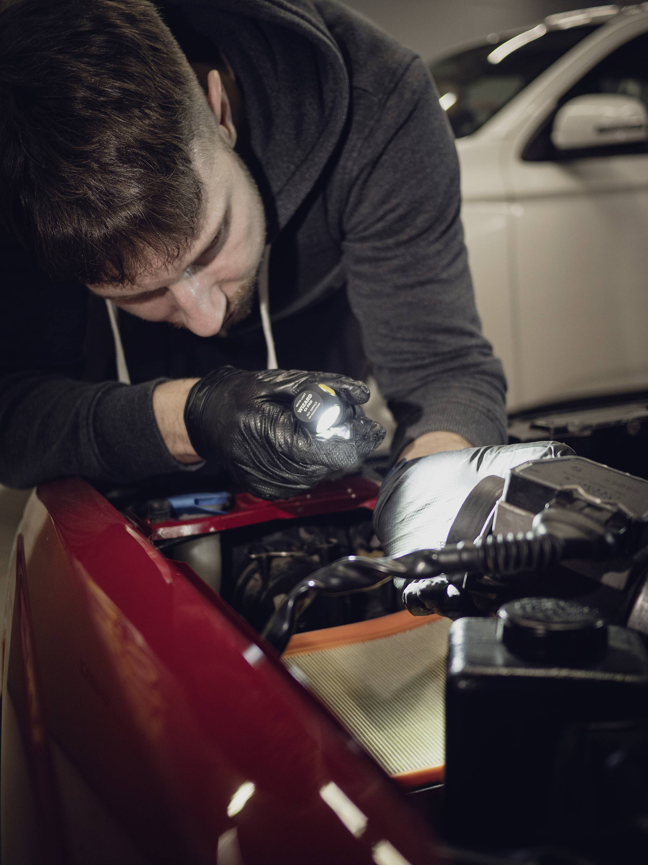 Mehaničar pregledava motor u radionici. Nosi crne rukavice i koristi lampu za inspekciju. U pozadini se vidi bijeli automobil.
