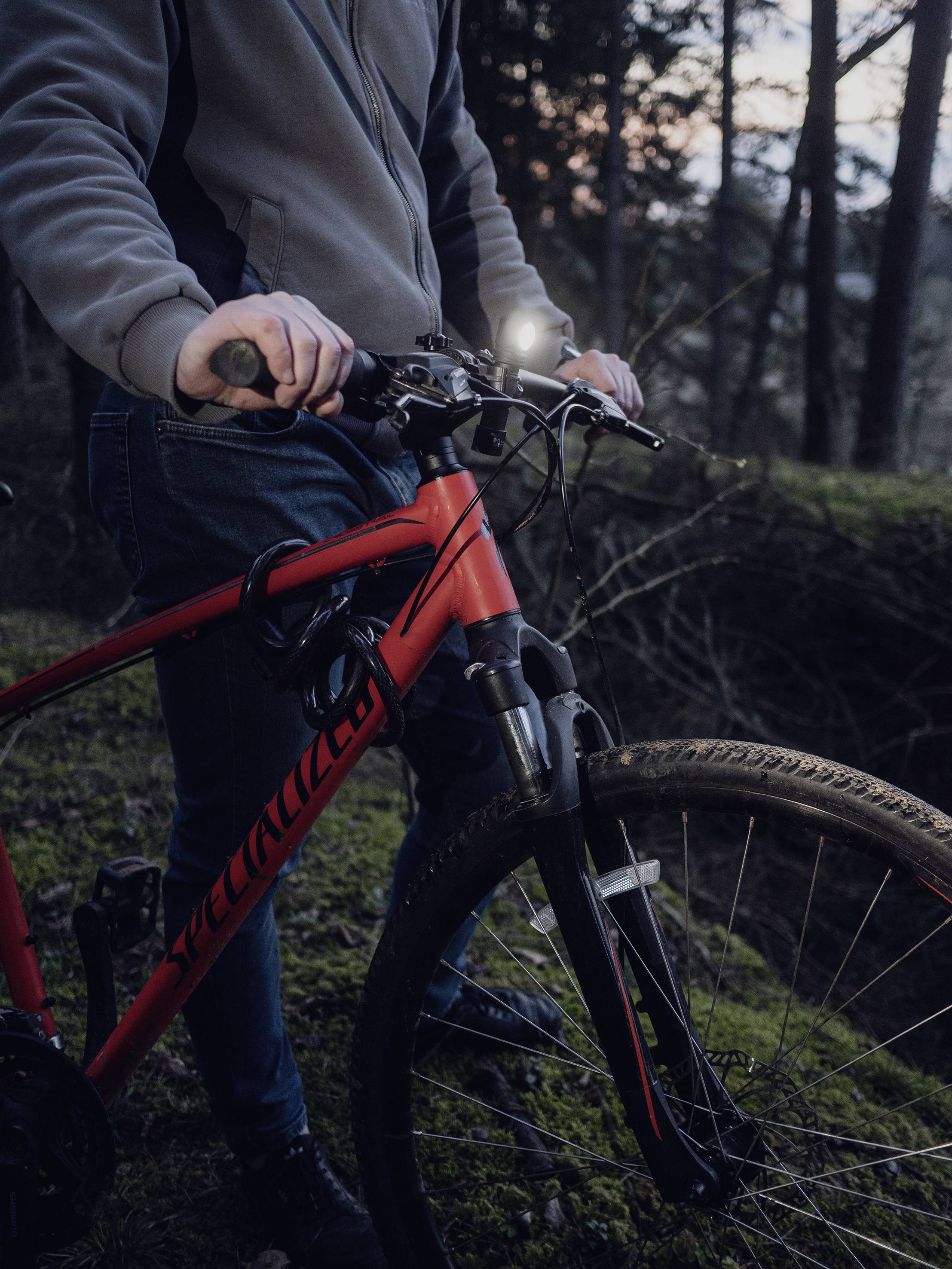 Osoba odjevena u outdoor odjeću drži crveni mountainbike na šumskom putu u sumrak.