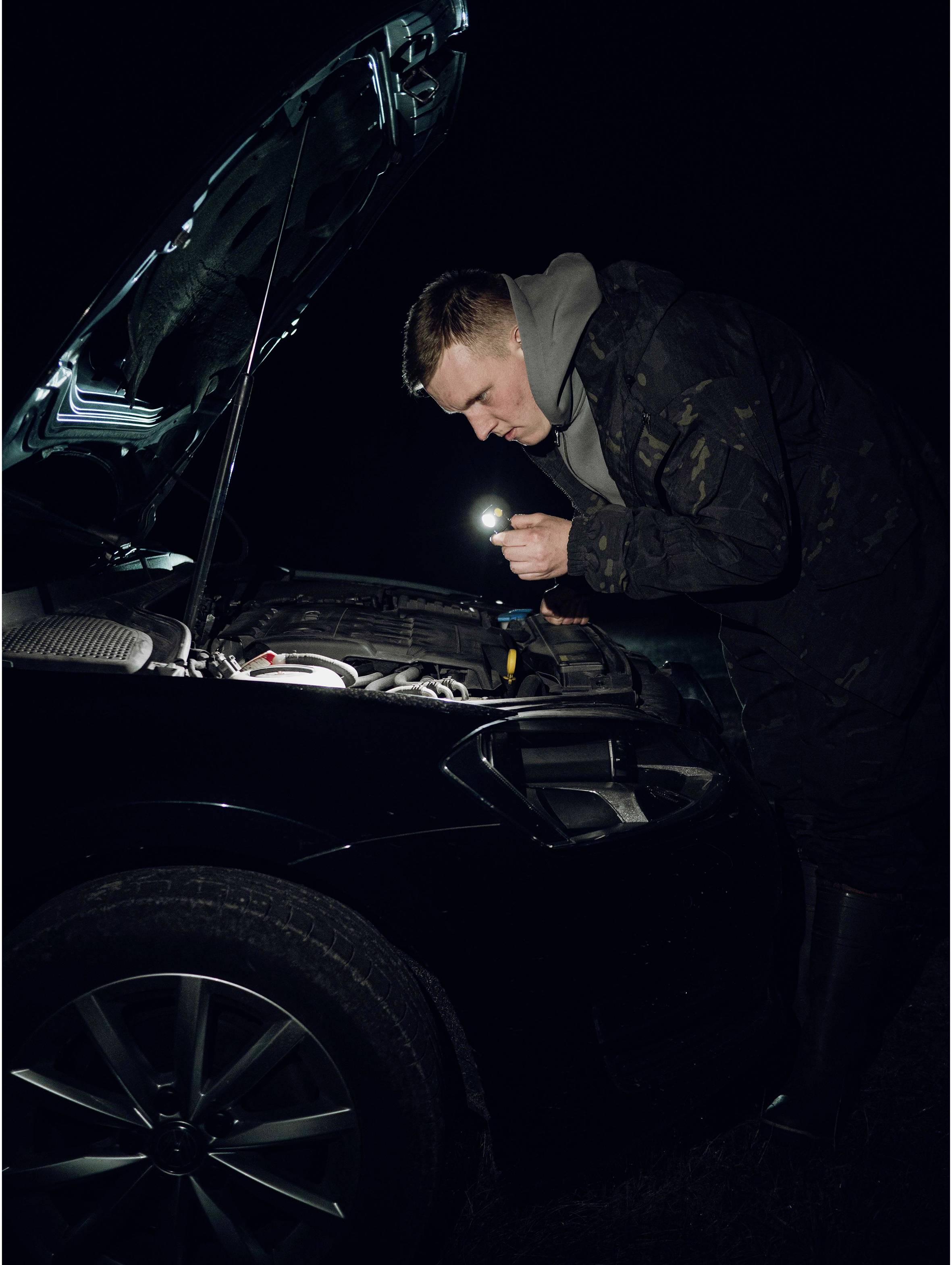 Muškarac provjerava motor automobila džepnom lampom tijekom noći. Poklopac motora je otvoren.