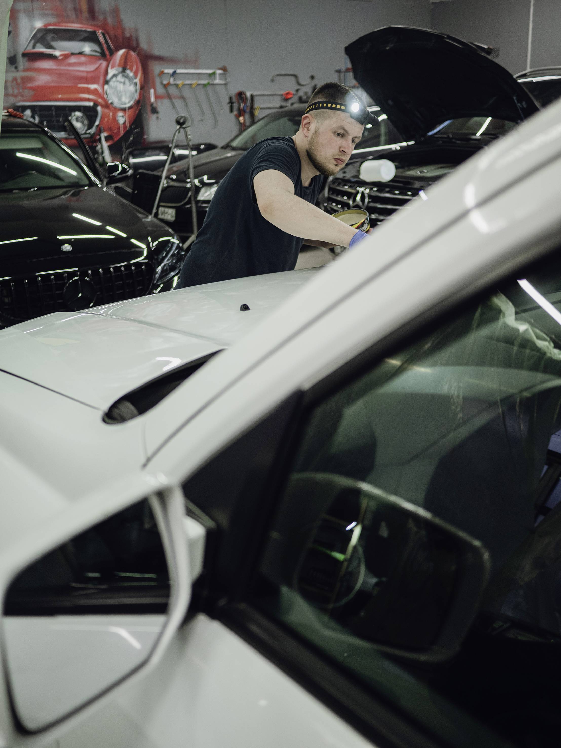 Muškarac popravlja automobil u radionici. Nosi čeonu lampicu i koncentrirano radi na prednjoj strani vozila.