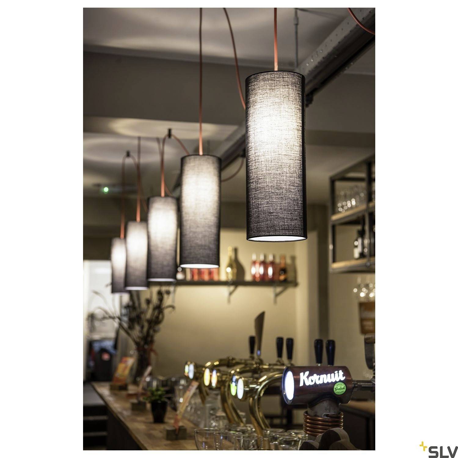 Viseće tkaninske lampe osvjetljavaju bar s točionicama i dekorativnim elementima. Ugodna atmosfera s toplim svjetlom.