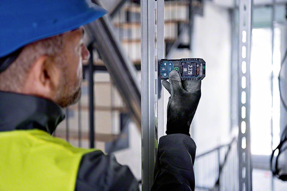 Građevinski radnik u zaštitnoj kacigi i radnom prsluku mjeri udaljenost mjernim uređajem u industrijskom okruženju.