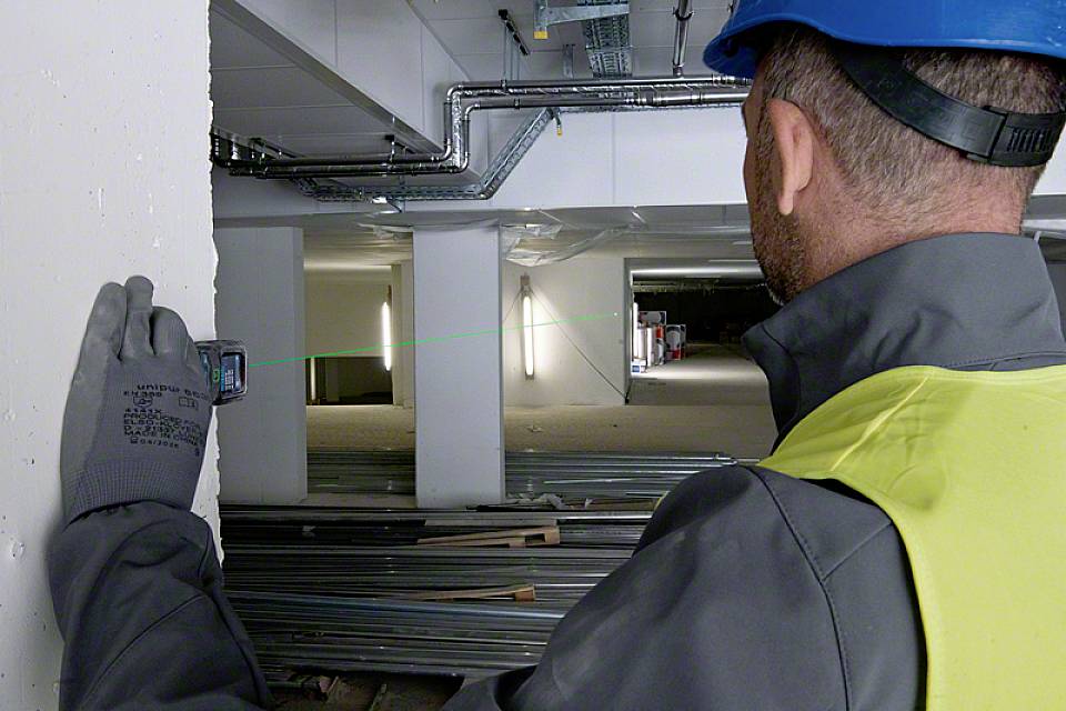 Građevinski radnik u zaštitnoj opremi mjeri udaljenost laserskim uređajem u nedovršenoj zgradi. Okruženje gradilišta s metalnim nosačima.
