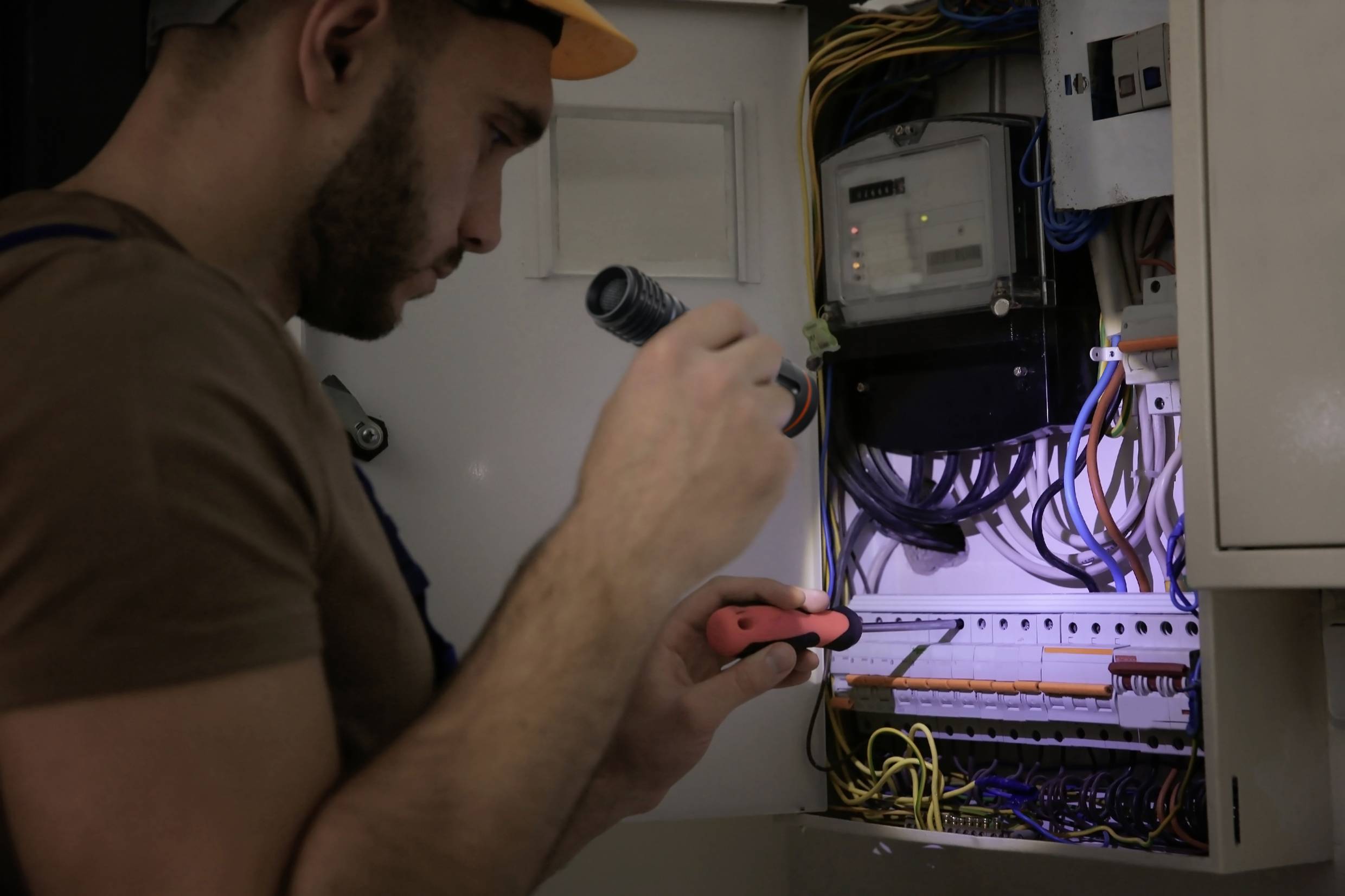 Električar radi u razvodnoj kutiji, drži džepnu lampu i koristi odvijač kako bi provjerio priključke.