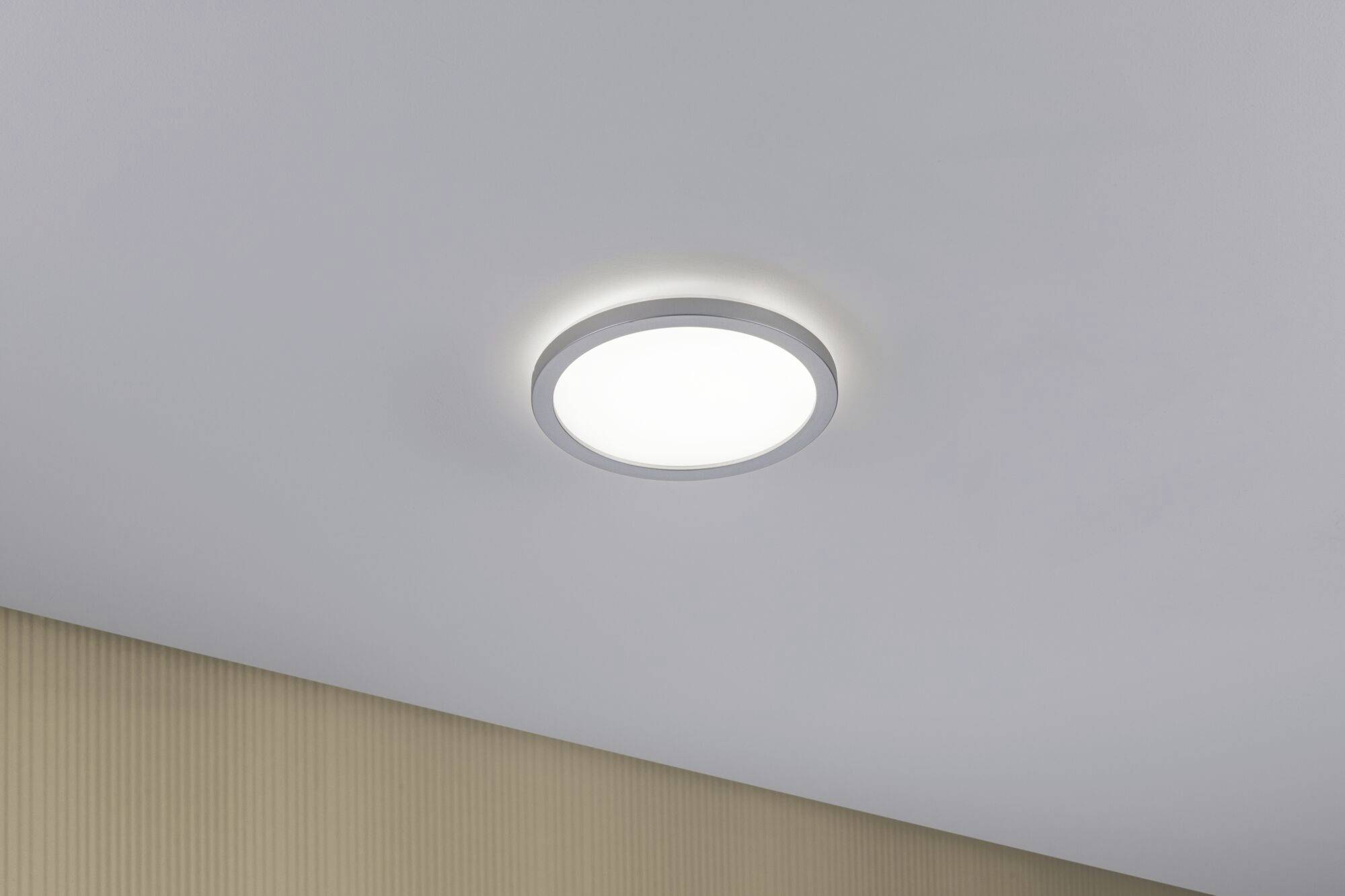 Okrugla stropna lampa na svijetlom stropu, osvjetljava prostoriju meko i ravnomjerno. Zidovi u pozadini su diskretno obojeni.