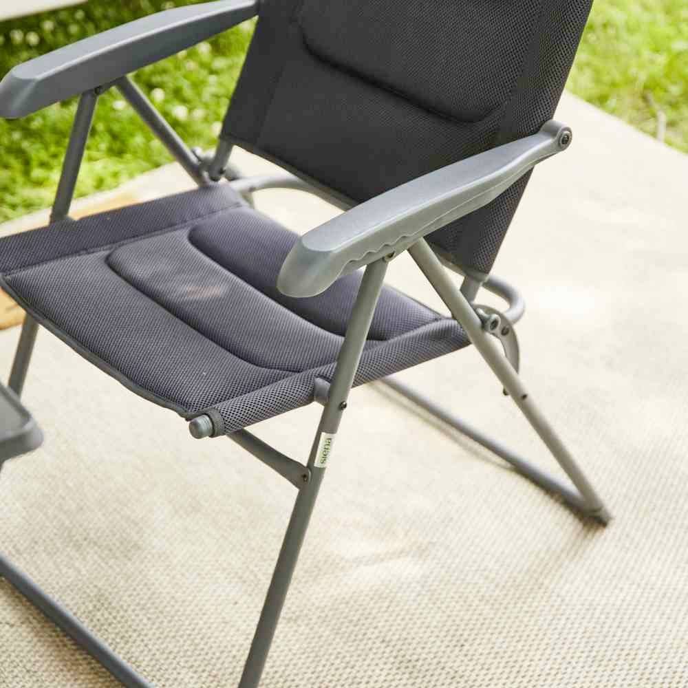 Crni sklopivi stolac stoji na tepihu, okružen zelenom travom vani. Stolac ima naslone za ruke i rešetkasti uzorak.