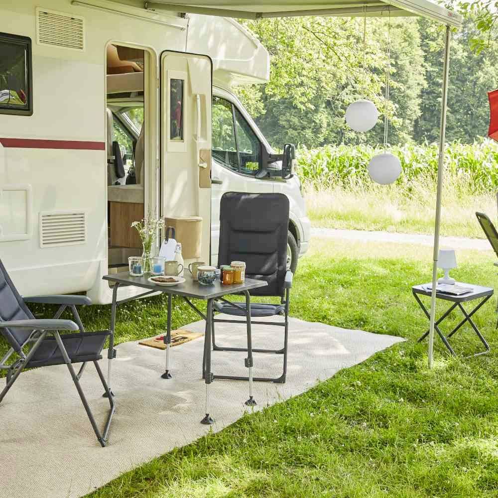 Kućni kamper na kamping prostoru s postavljenim stolom, stolcima i posuđem u zelenom okolišu.