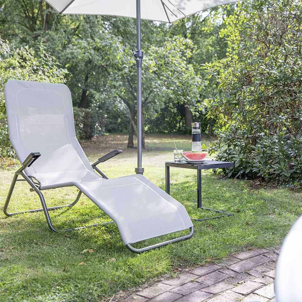 Bijela ležaljka za sunčanje stoji u vrtu ispod otvorenog suncobrana. Pokraj nje je mali stolić s karafi vodom i tanjurom s lubenicom.