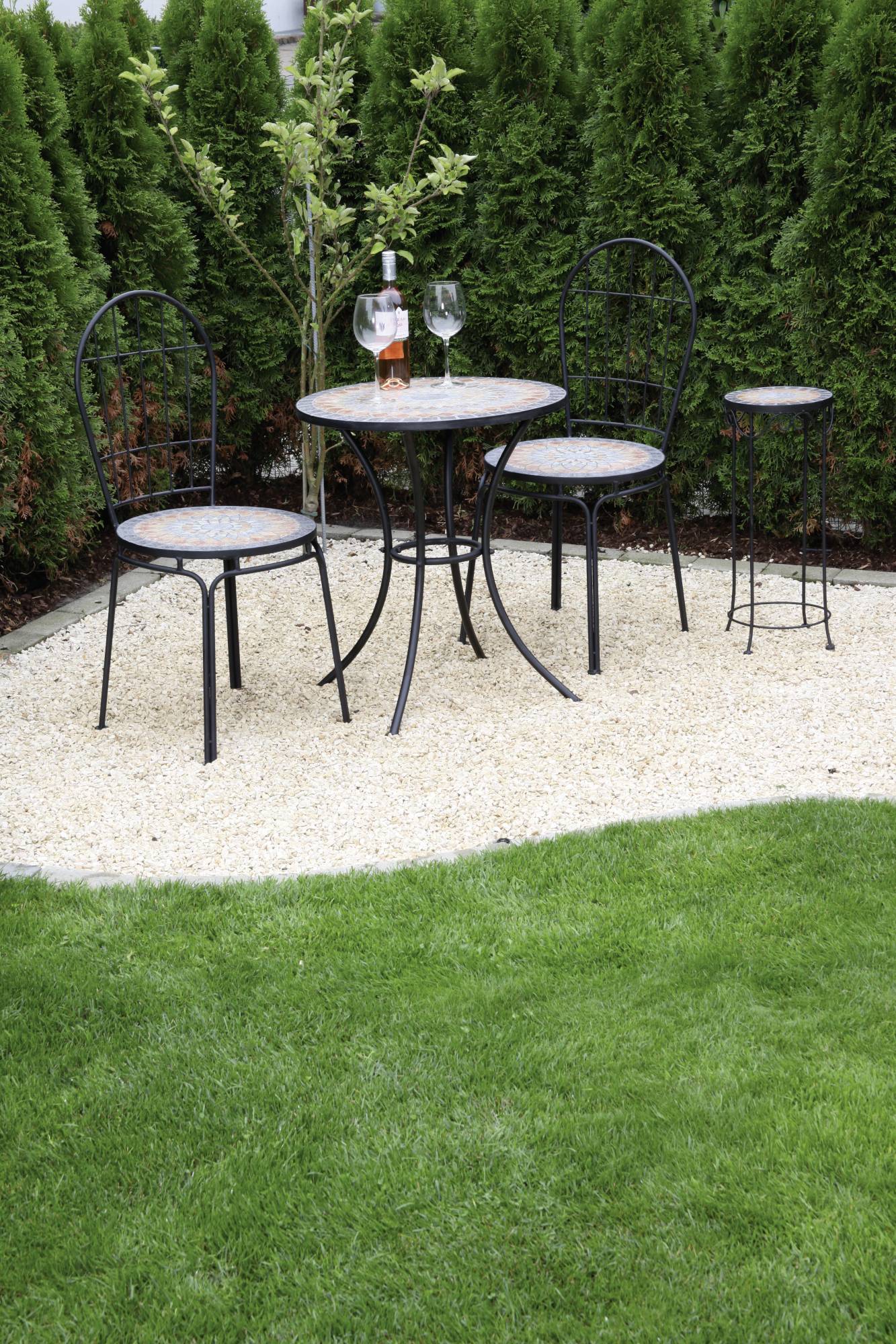 Mali vrtni stolić s dvije stolice na šljunčanom tlu, okružen zelenim biljem. Na stolu stoje boca i dvije čaše.