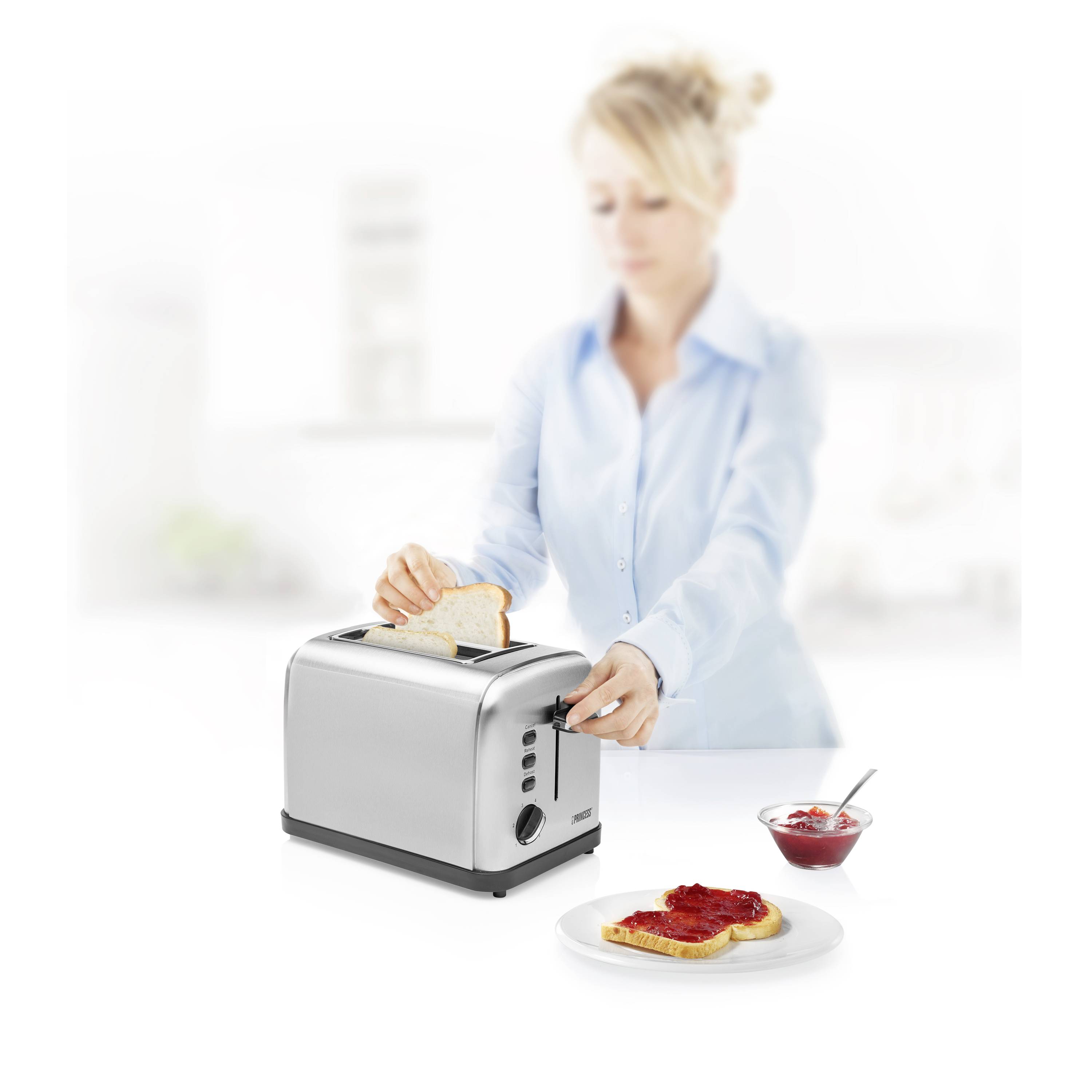 Žena stavlja tost u toster u svijetloj kuhinji. Ispred nje je stol s toastom i džemom od jagoda.