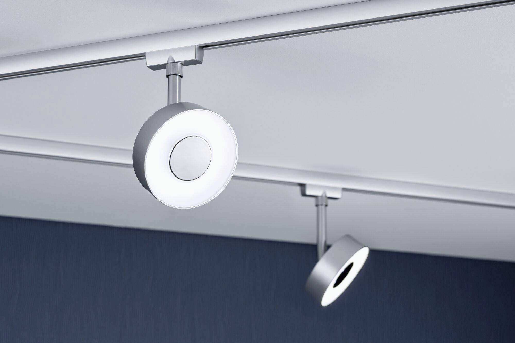 Dvije moderne, okrugle stropne lampe u srebrnastoj boji pričvršćene su na šini i svjetle prema dolje. Vise na elegantnom plavom stropu.