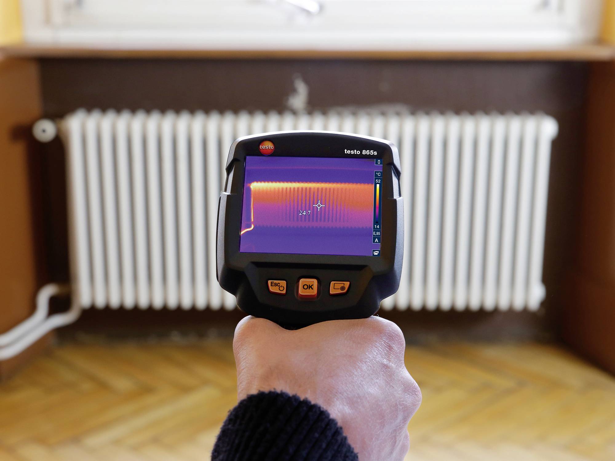 Jedna ruka drži termičku kameru koja prikazuje raspored temperature grijača. Zaslon pokazuje tople i hladne zone.