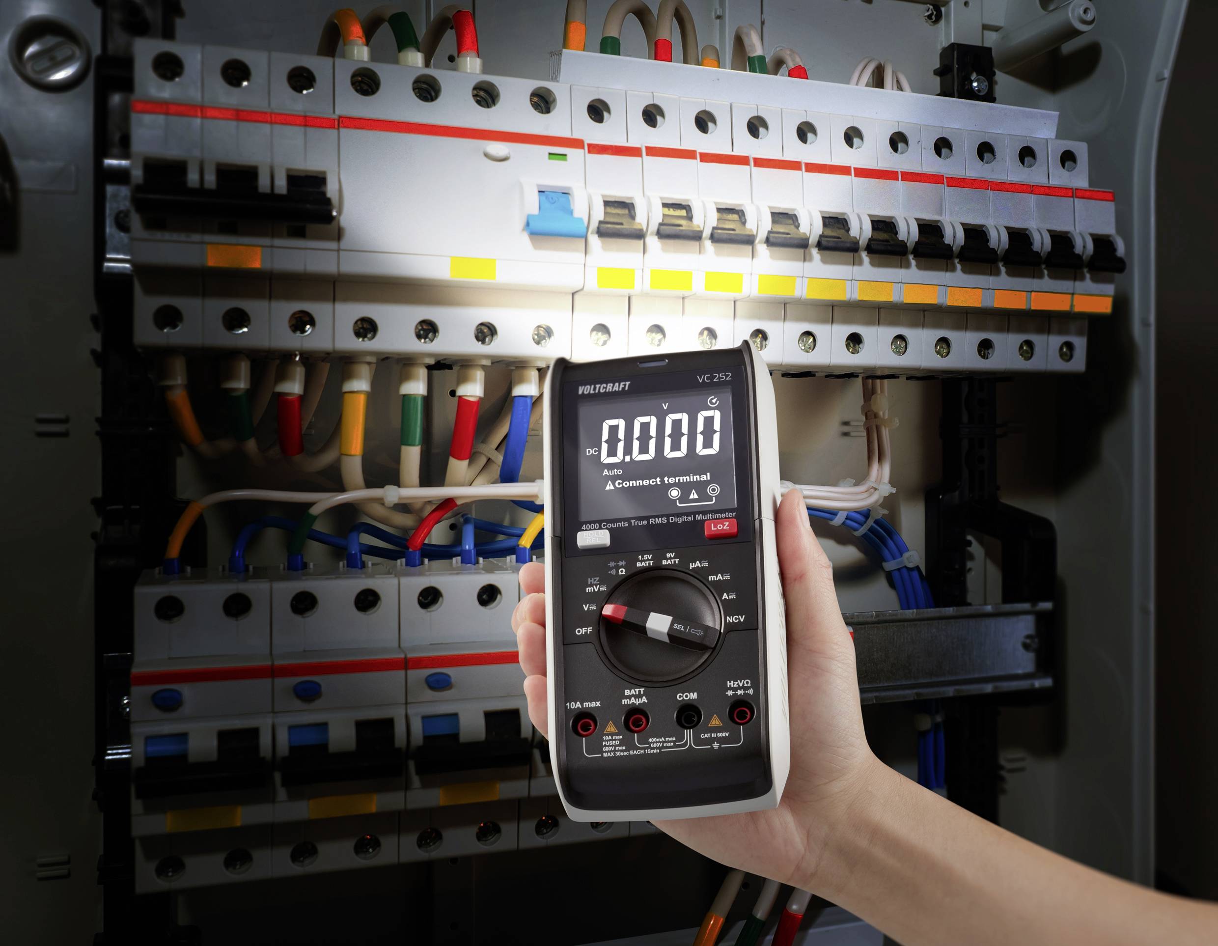 Jedna ruka drži digitalni multimetar koji pokazuje napon od 0,000 ispred otvorene električne razvodne kutije s nekoliko kabela.