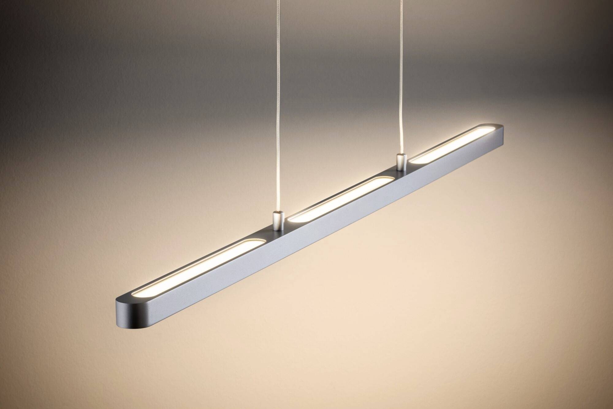 Moderna, uska viseća lampa s dva kabela koja ravnomjerno emitira toplo bijelo svjetlo. Visi horizontalno u minimalistički uređenoj prostoriji.