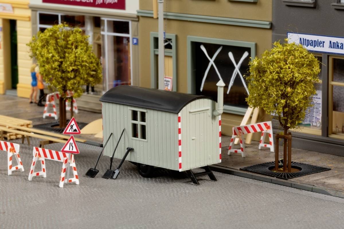 Kiosk na kotačima stoji na gradskoj ulici, okružen crveno-bijelim barijerama i dva stabla. U pozadini su vidljive fasade zgrada.