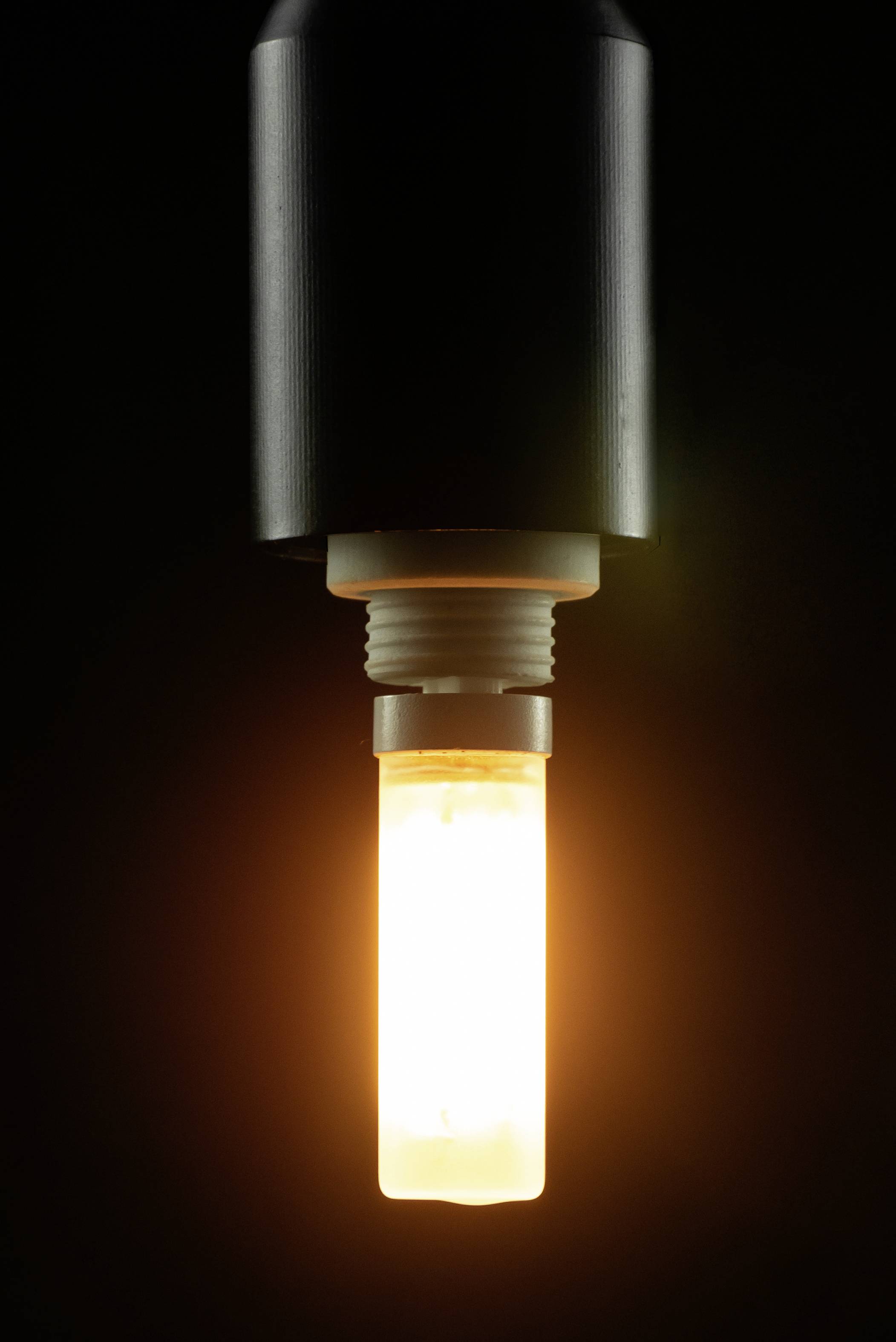 Sjajnja žarulja visi s crnog nosača, pred tamnom pozadinom. Lampa emitira toplo, narančasto svjetlo.