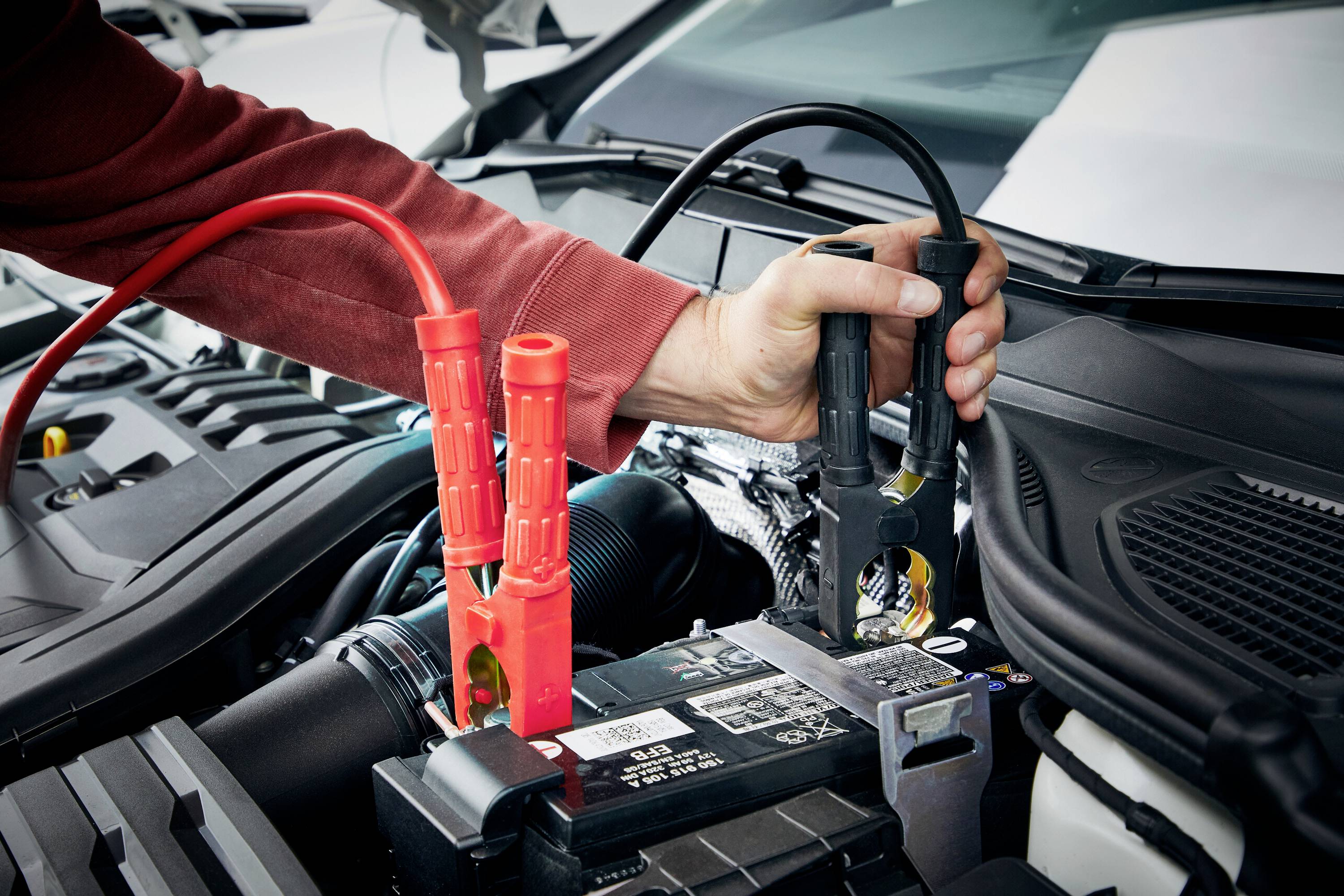 Jedna ruka spaja pogonske kablove s automobilskom baterijom kako bi pokrenula automobil. Crveni kabel je spojen s pozitivnim polom.