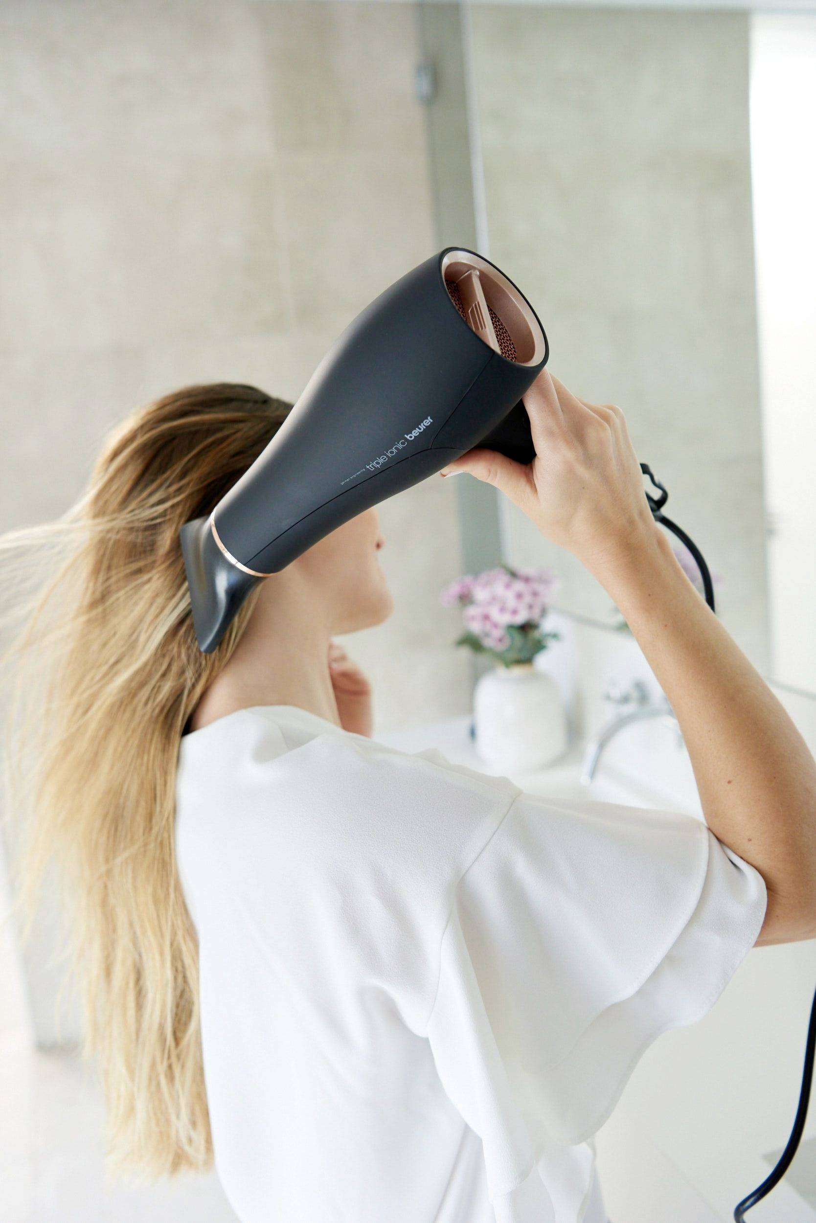 Žena suši kosu sušilom za kosu u kupaonici. U pozadini se na umivalniku nalazi vaza s ružičastim cvjetovima.