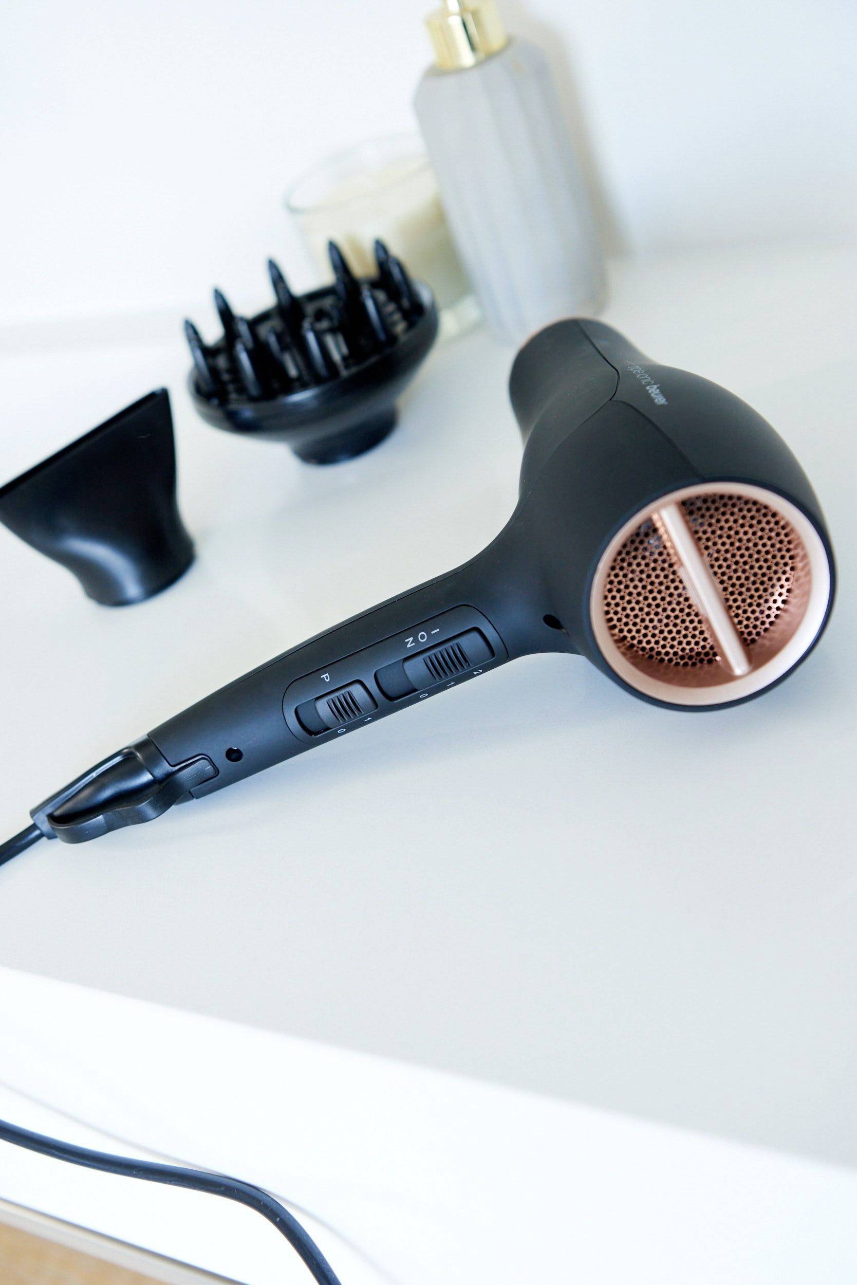Crni sušač za kosu s različitim nastavcima leži na bijelom stolu. U pozadini se nalaze svijeća i boca.