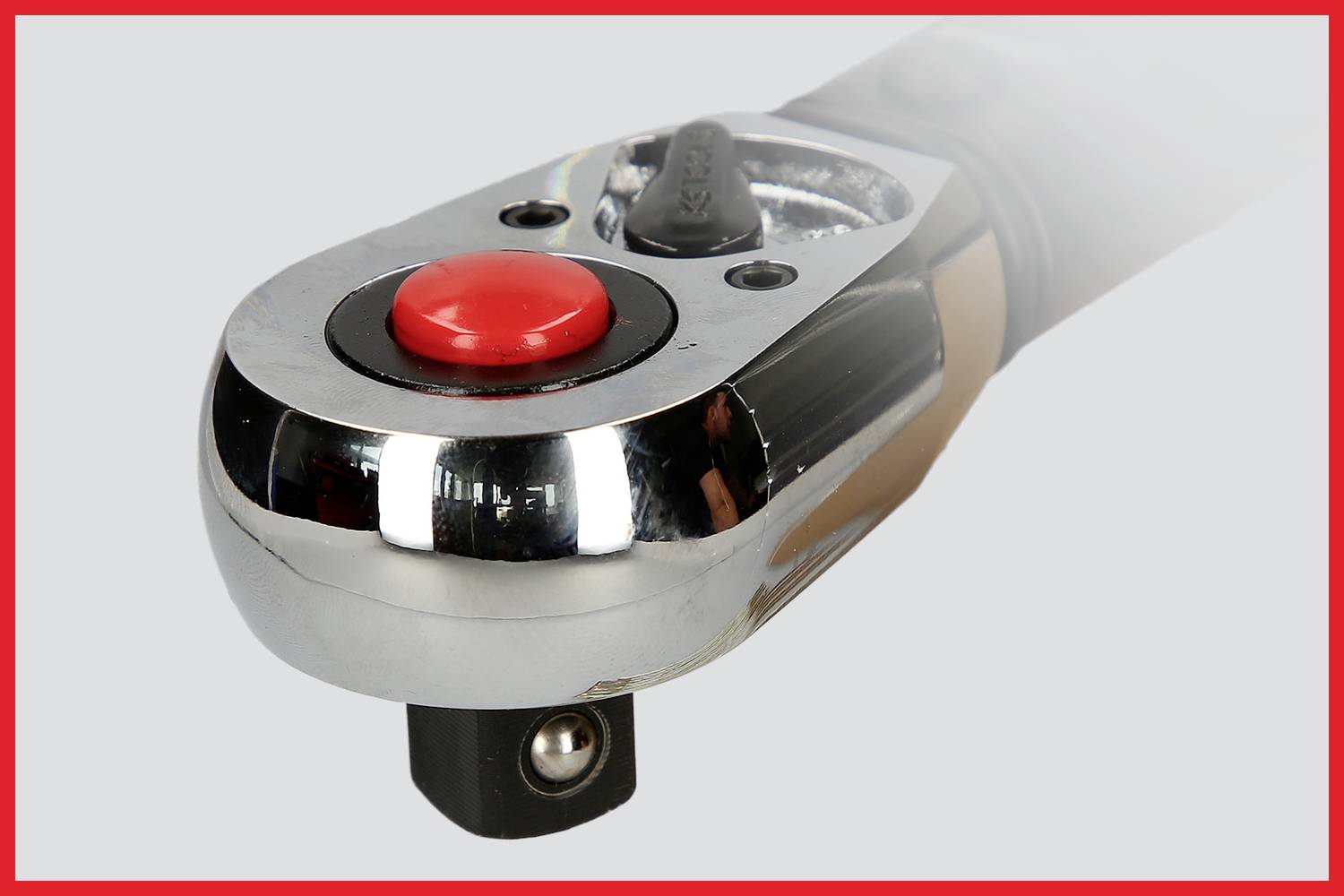 Bliski kadar kromiranog alata s crvenim gumbom, vjerojatno ručke momentnog ključa, na sivoj pozadini.