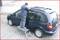 Muškarac stoji na ljestvama pored plavog automobila i radi na krovu, na popločenom tlu ispred zgrade.
