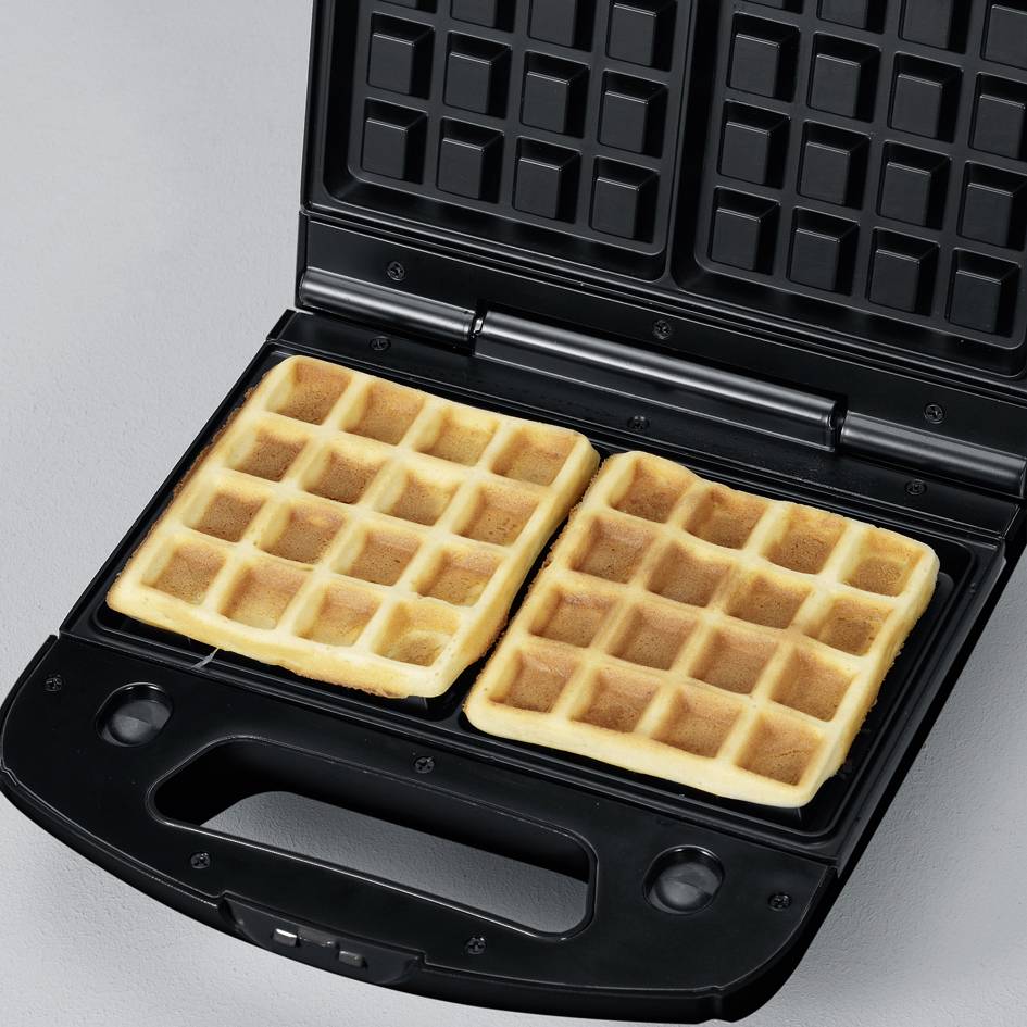 Dvije pečene waffle u otvorenom aparatu za waffle na sivoj podlozi.
