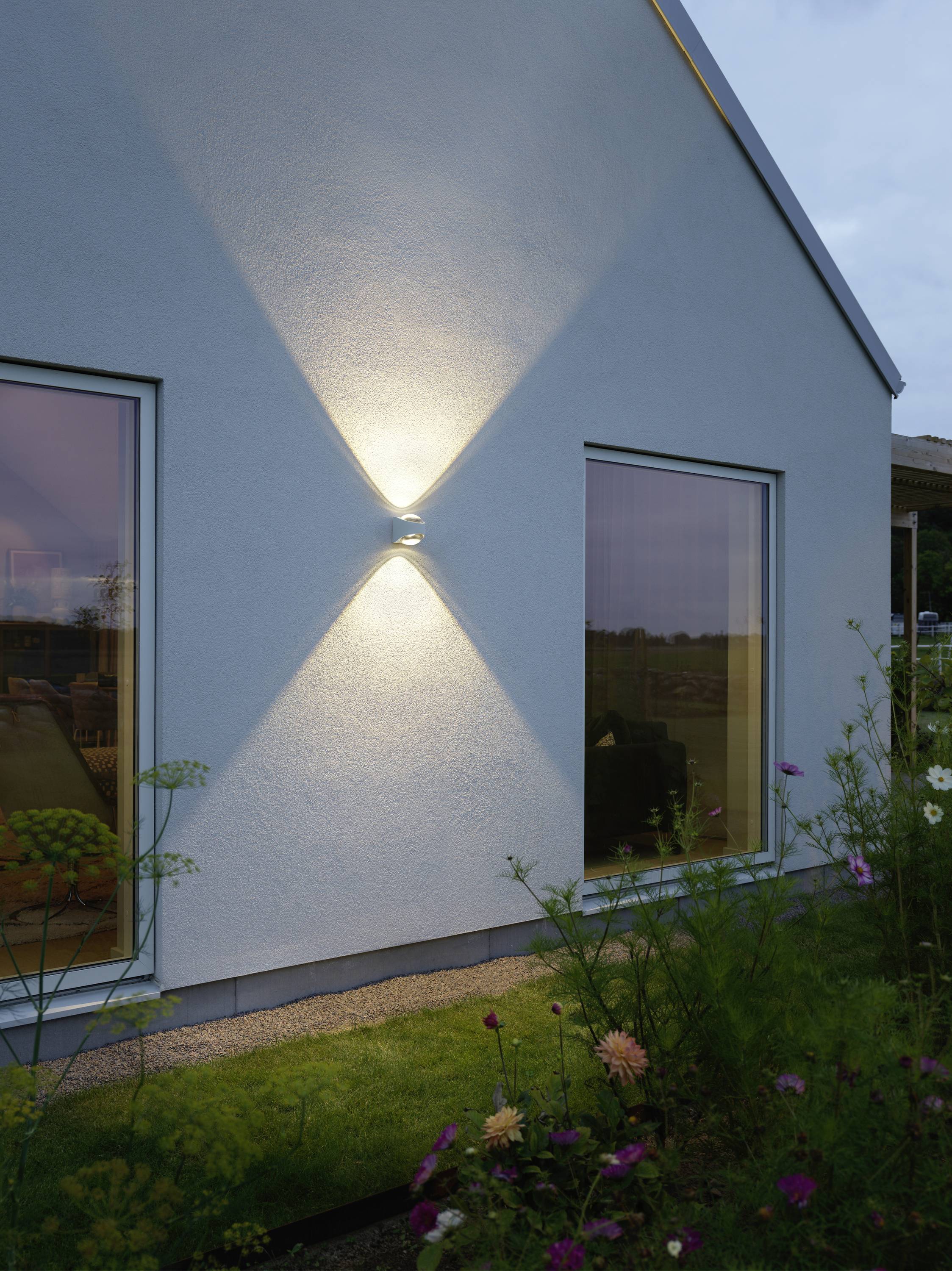 Moderna kuća s osvjetljenim vanjskim zidom u sumrak, okružena vrtom punim cvijeća, prenosi mirnu atmosferu.