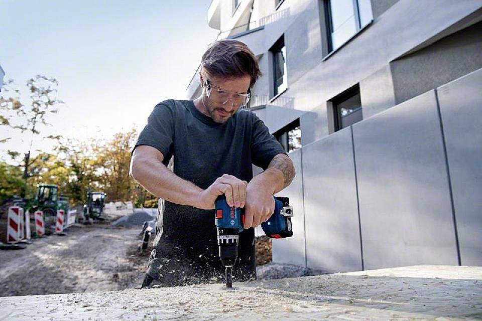 Muškarac buši drvenu dasku električnom bušilicom na gradilištu. U pozadini se naziru građevinski strojevi.