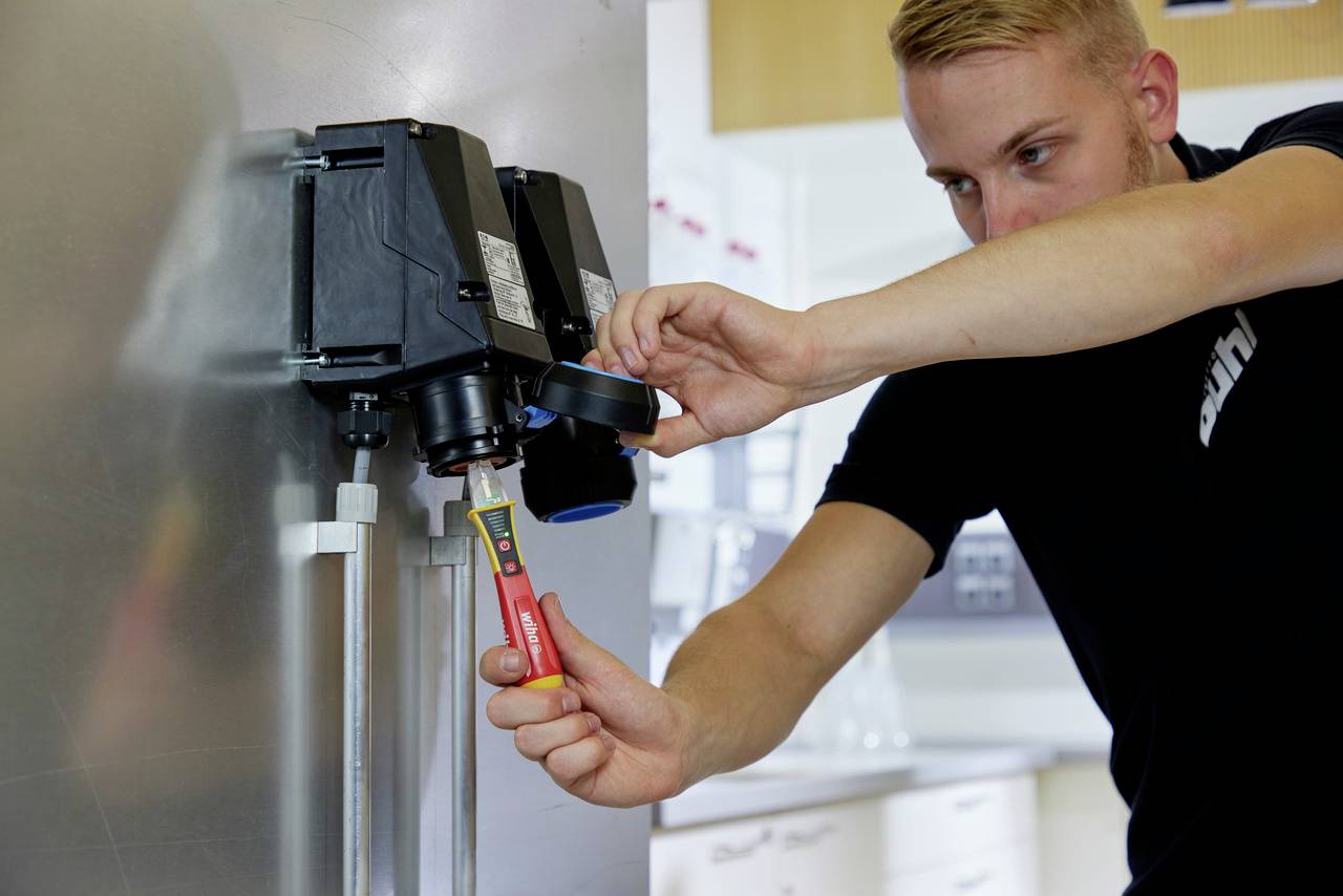 Muškarac radi na elektroničkoj jedinici pomoću odvijača. Nosi crnu majicu i stoji u tehničkoj prostoriji.