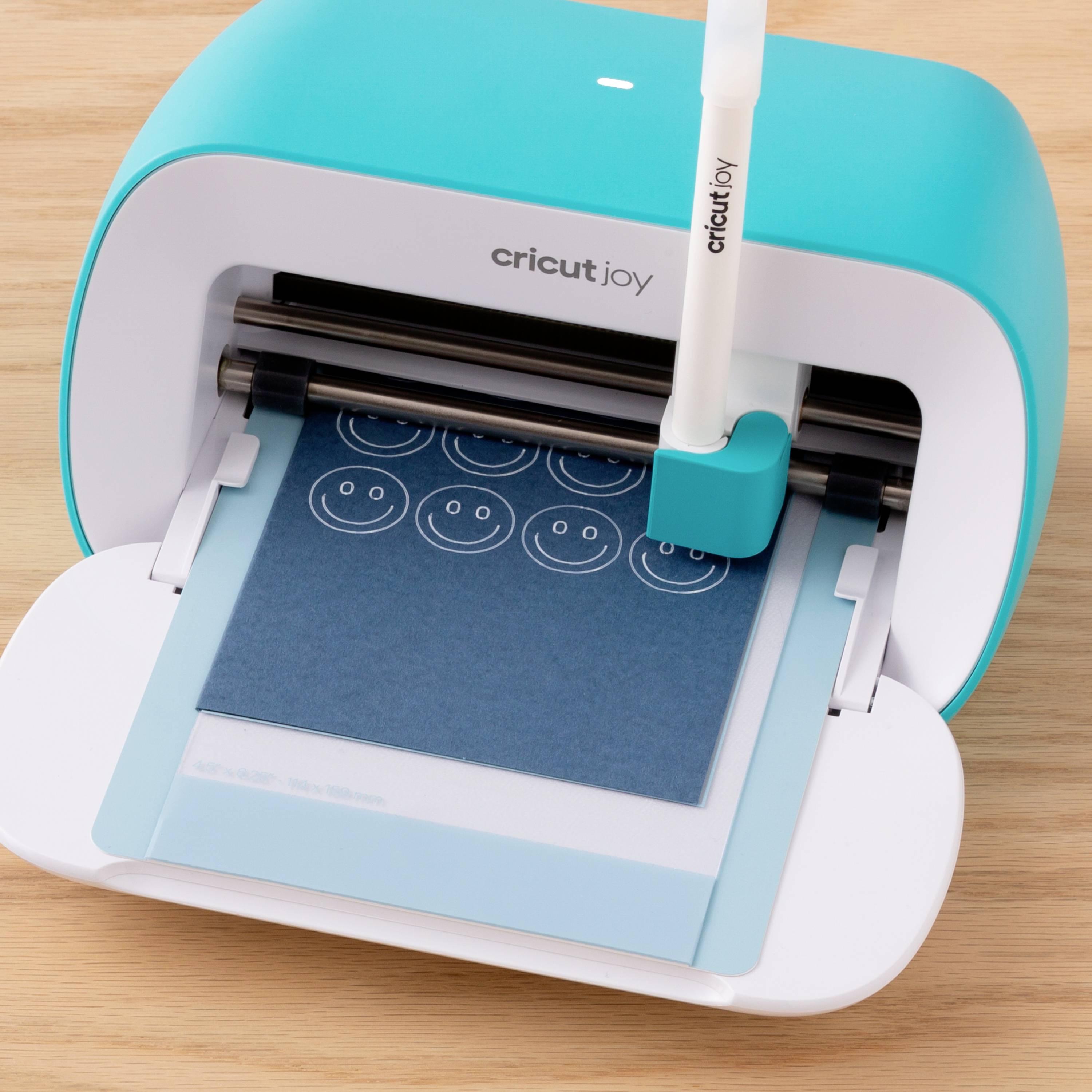 Cricut Joy stroj za rezanje koji izrezuje plavi karton sa više smiley lica, stoji na drvenom stolu.