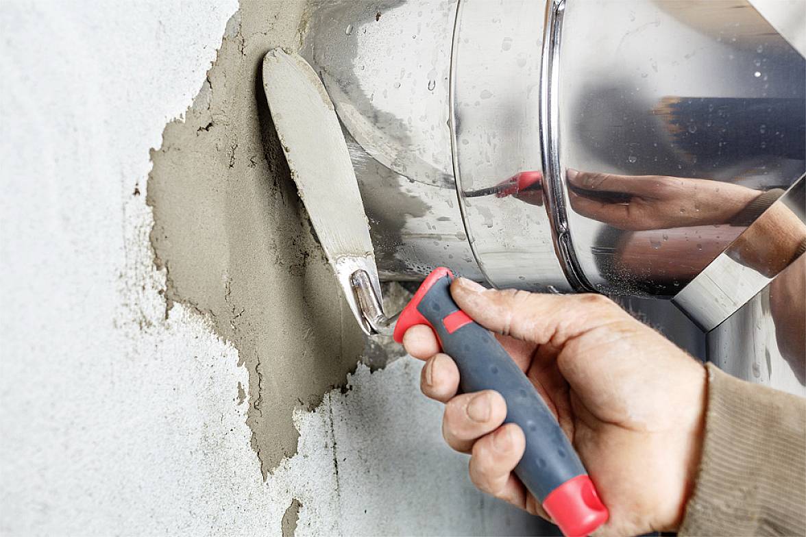 Jedna ruka spatulom nanosi beton na zid. U pozadini srebrni mješač.