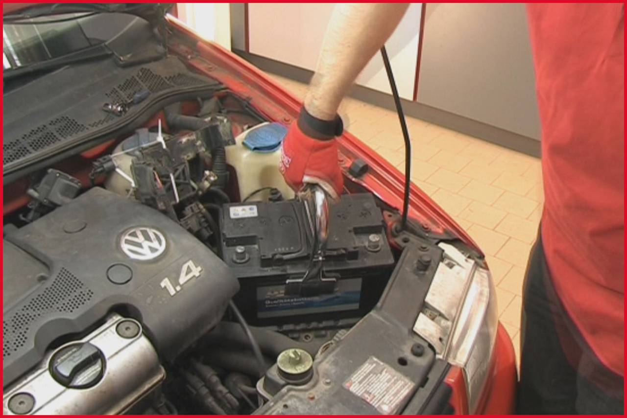 Mehaničar provjerava automobilsku bateriju u crvenom vozilu. Jedna ruka drži dijagnostički alat na bateriji.