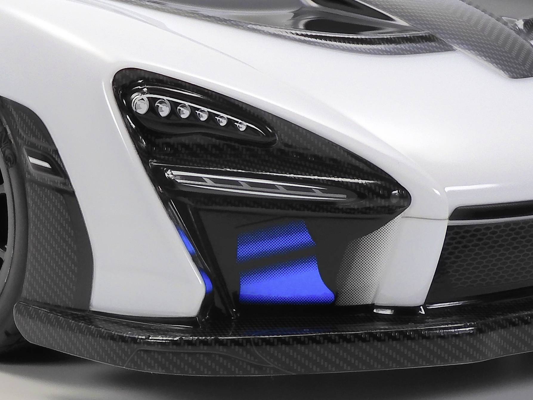 Detaljan pogled na bijeli automobil s karakterističnom prednjom maskom i plavim svjetlosnim elementima, s naglascima od crnog karbona.